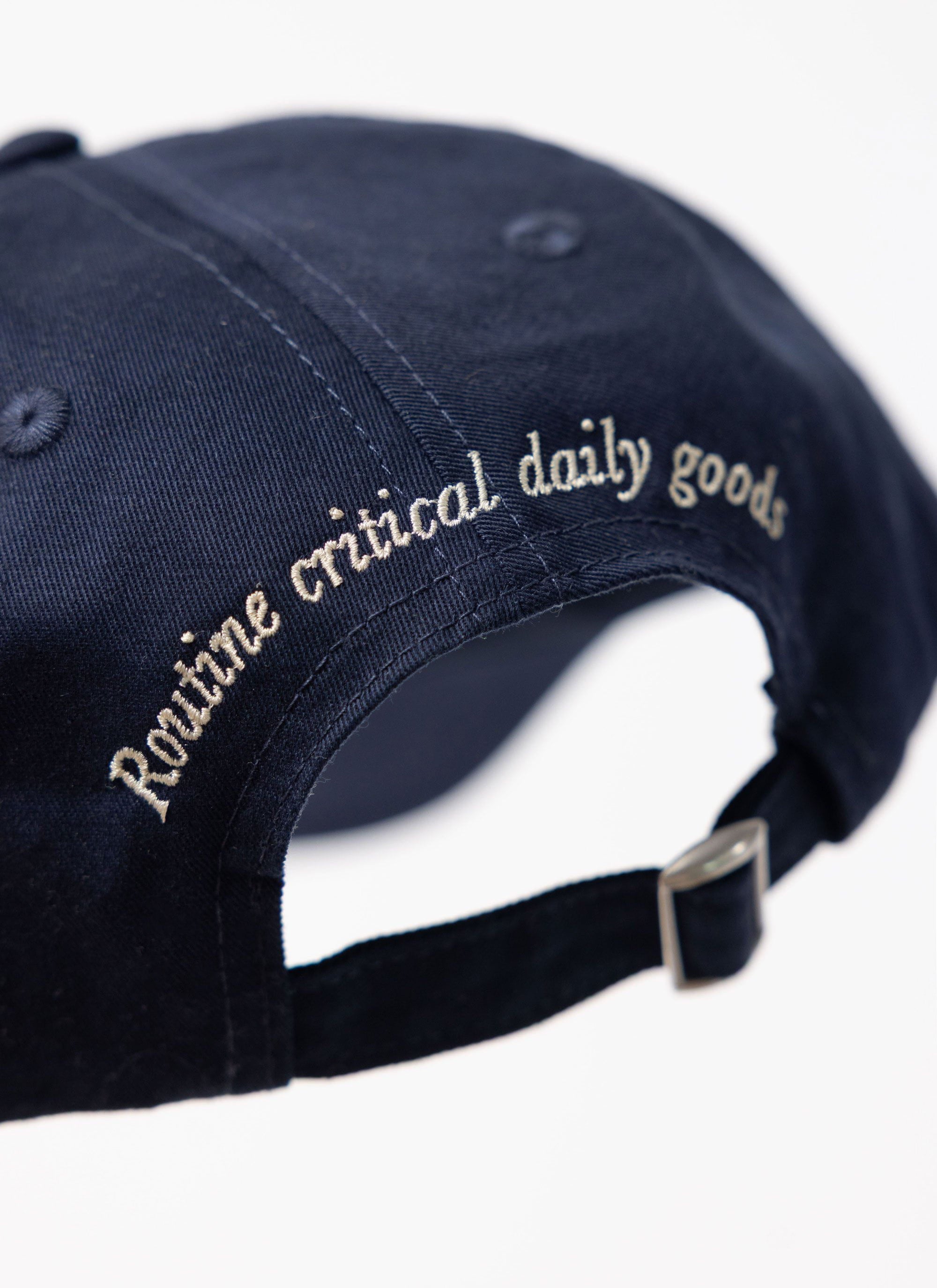 Close-up of the Percival World Key Cap in navy, detailing the 'Routine critical daily goods' embroidery on the back.