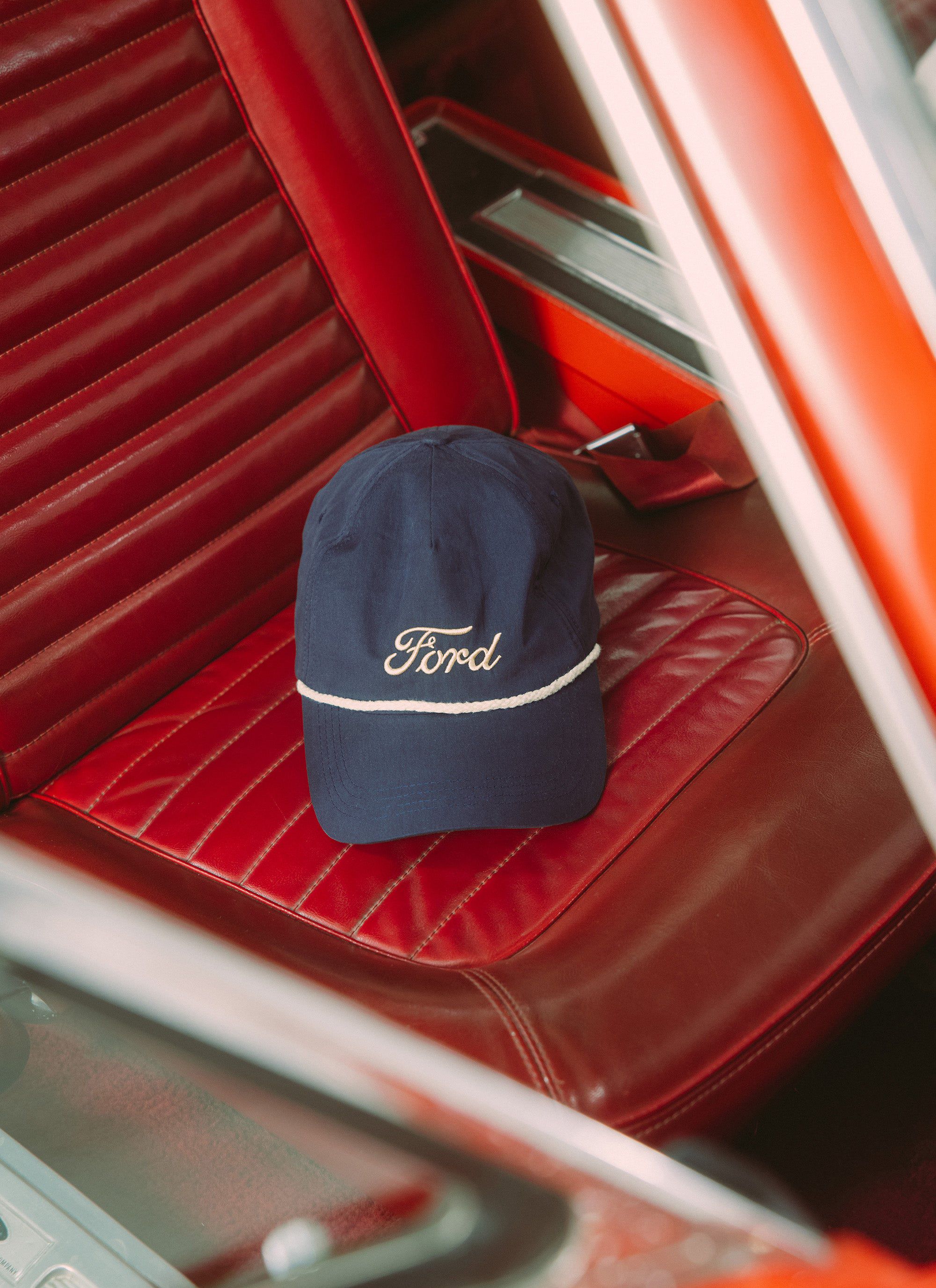 A navy Percival x Ford trucker cap with ecru embroidery sitting on the red leather seat of a classic Ford Mustang.