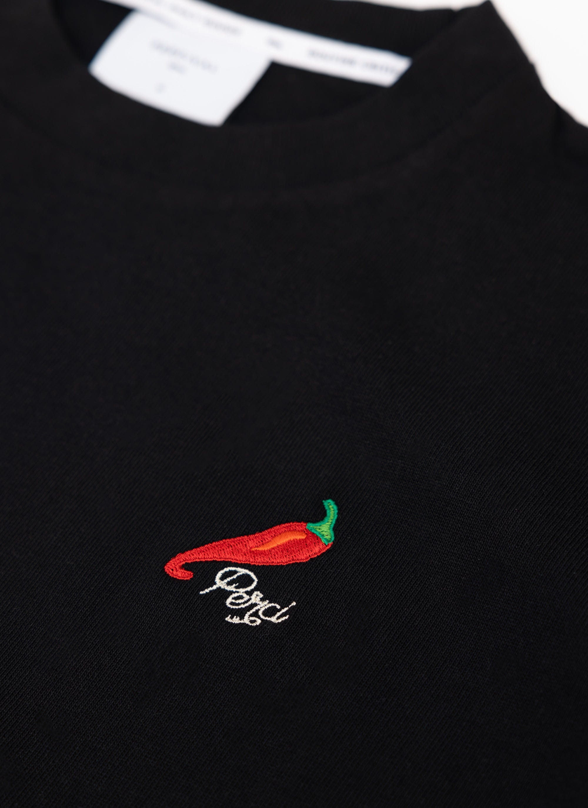 A close-up of the red chilli and logo embroidery on the chest of the black Percival Loyalty T Shirt.