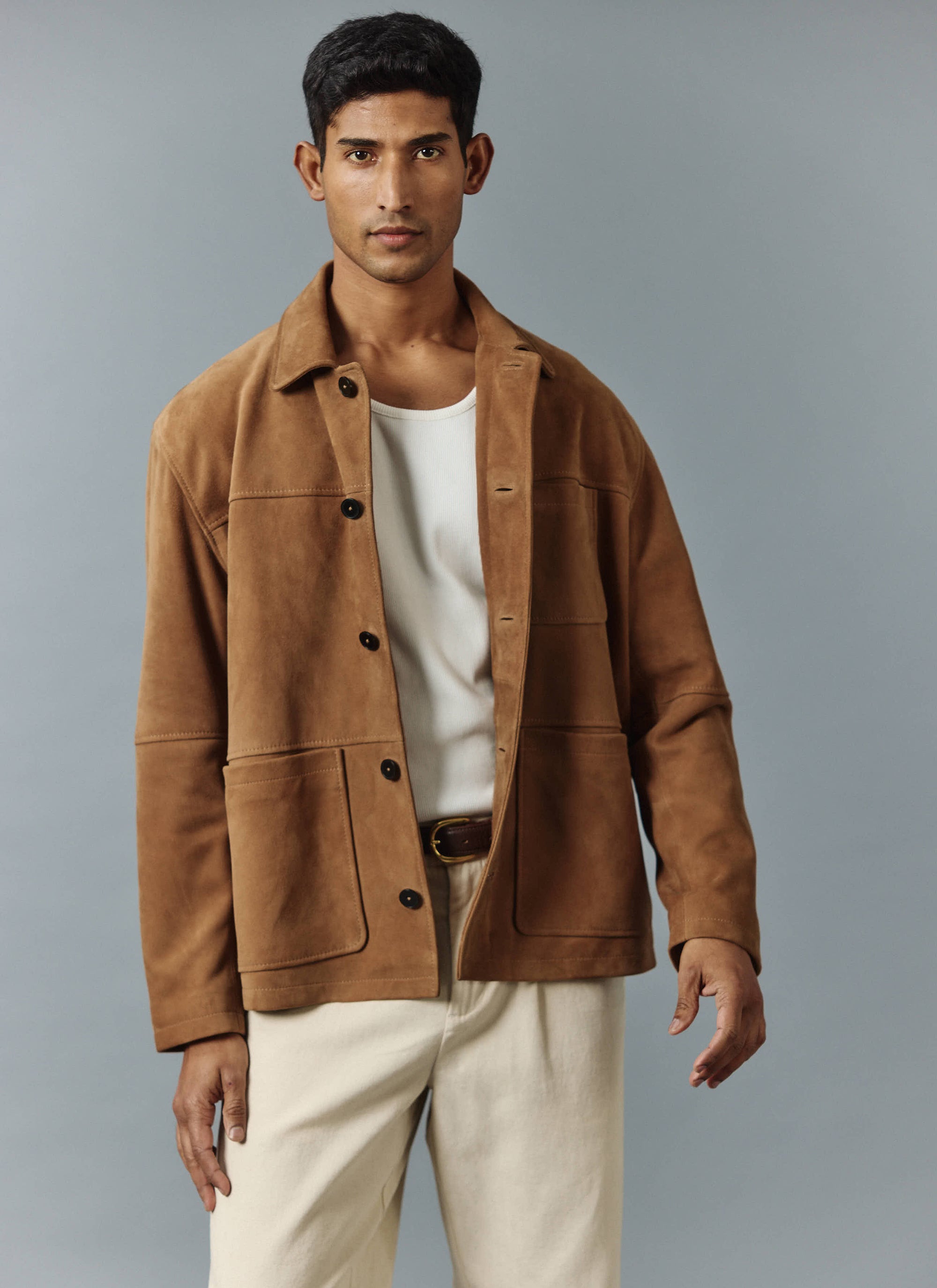 A model wears the Percival Afterward Suede Chore Jacket in tan, styled open over a white vest and cream trousers.
