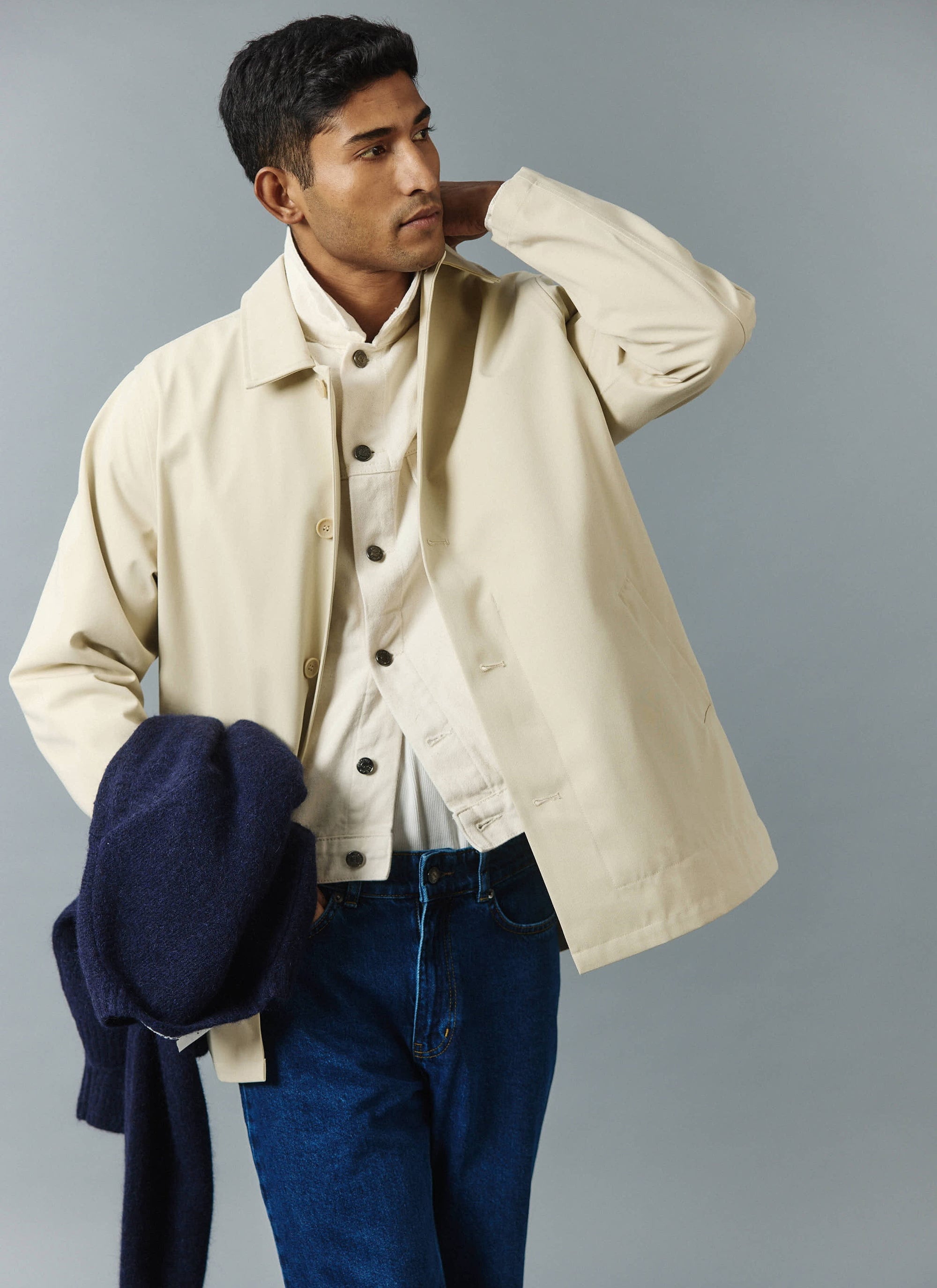 A model wears the Percival Sherlock Short Trench Coat in stone over a layered outfit, holding a navy jumper.
