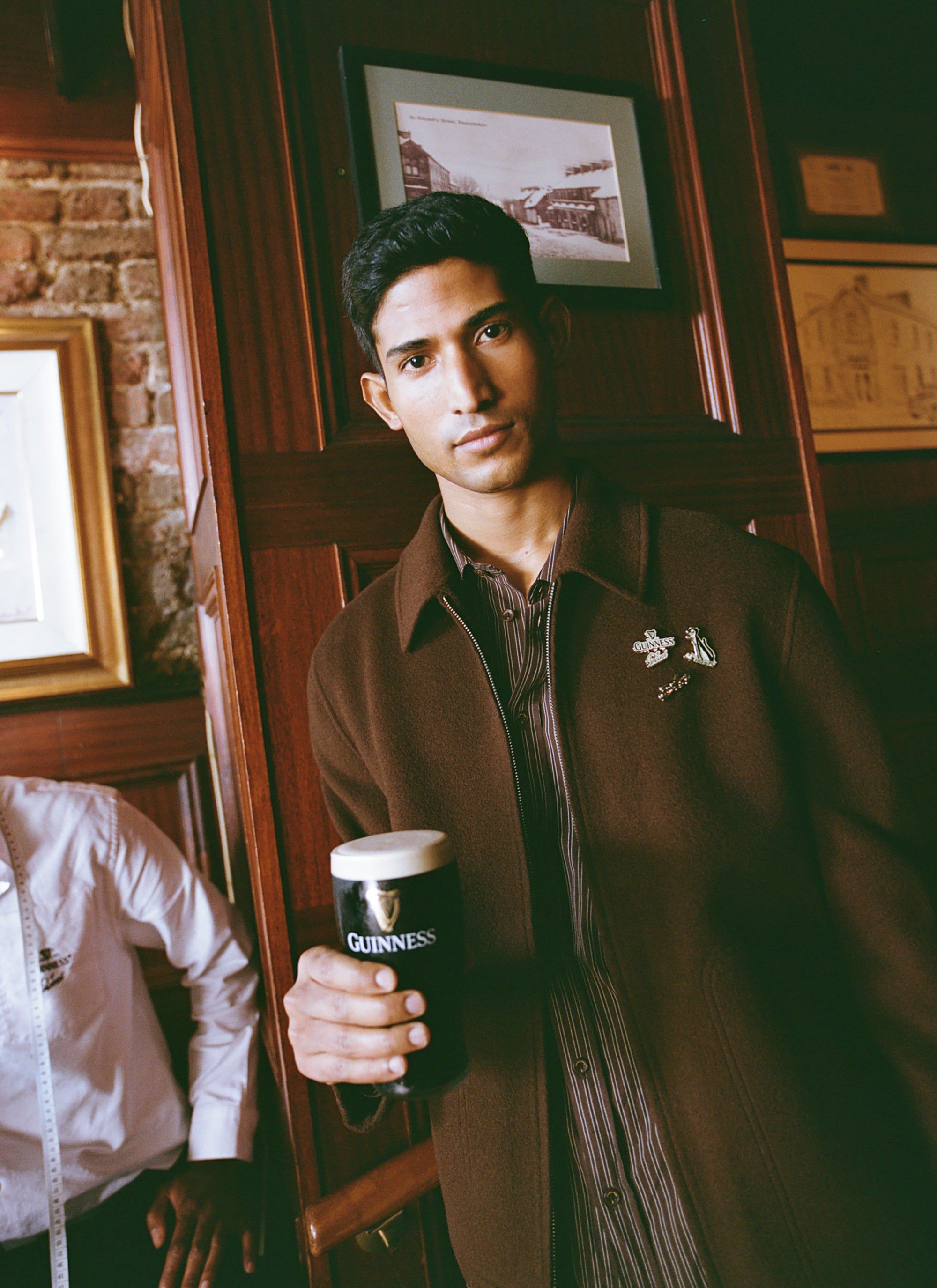 A model wears the Guinness x Percival Wool Jacket in espresso, holding a pint of Guinness in a classic pub setting.