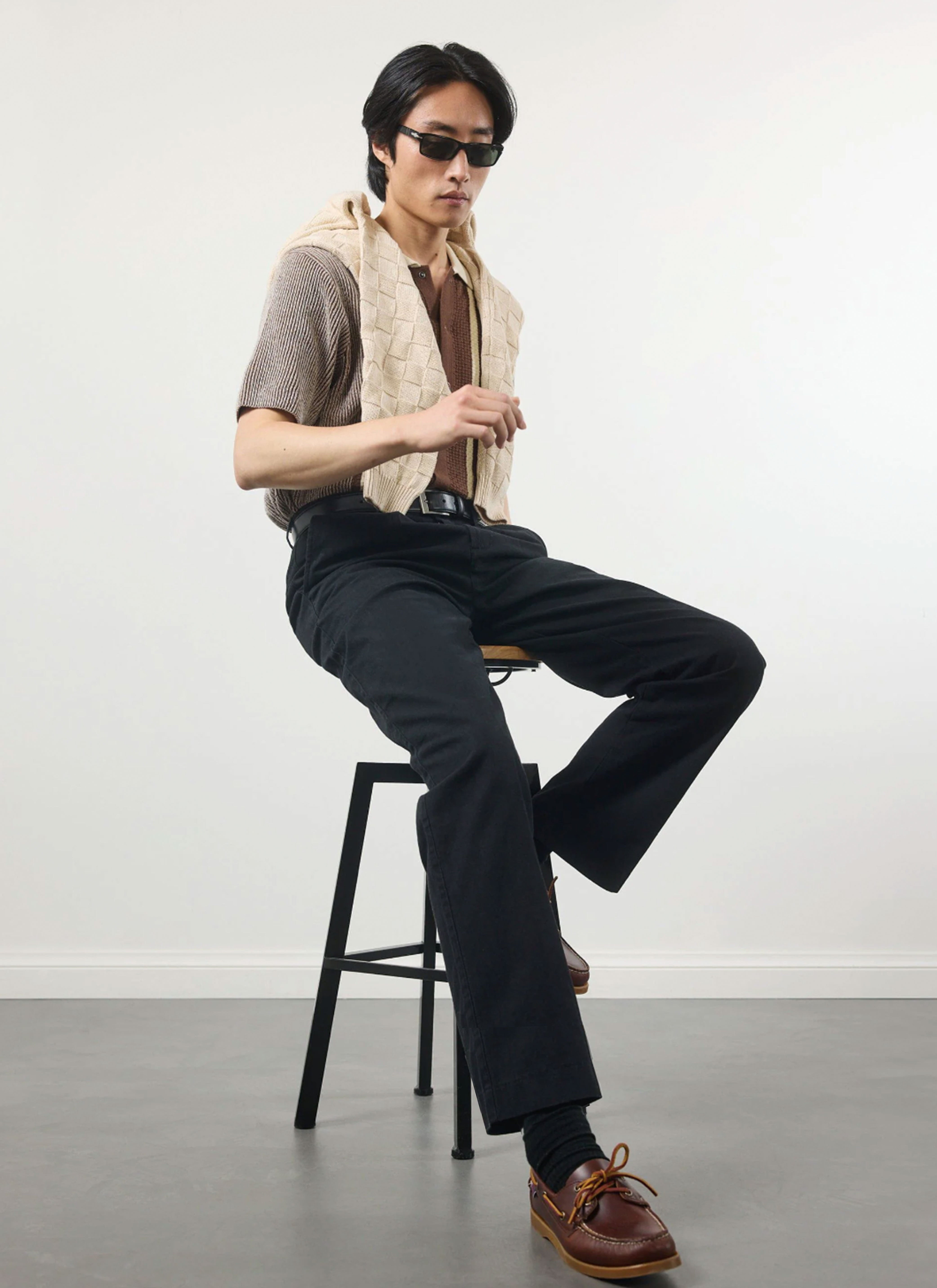 A model sits on a stool wearing the Percival Teramo Knitted Polo Shirt in ecru with a jumper draped over his shoulders.
