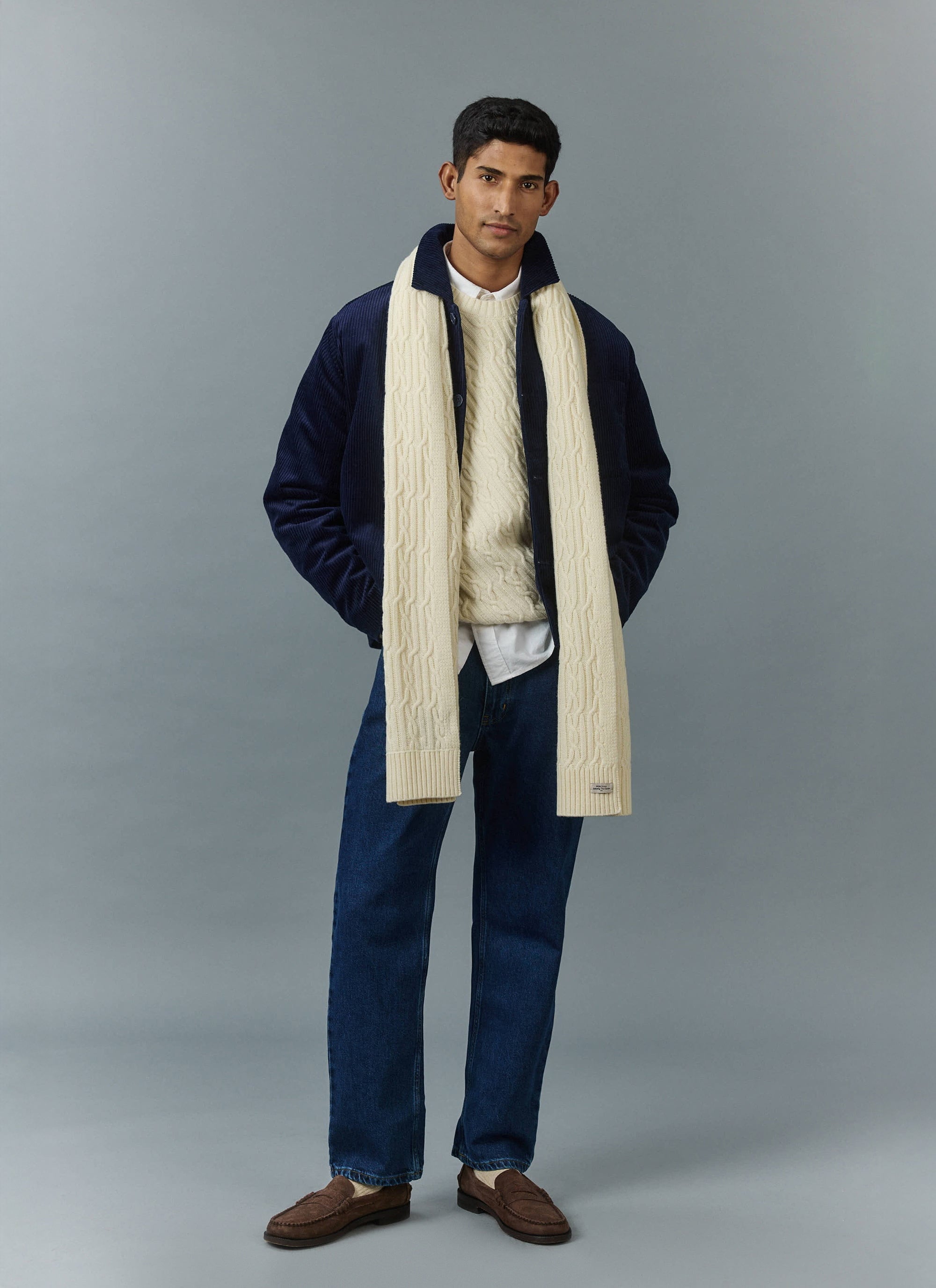 A model wears a Percival navy cord overshirt, ecru cable knit jumper and matching scarf, styled with dark denim jeans.