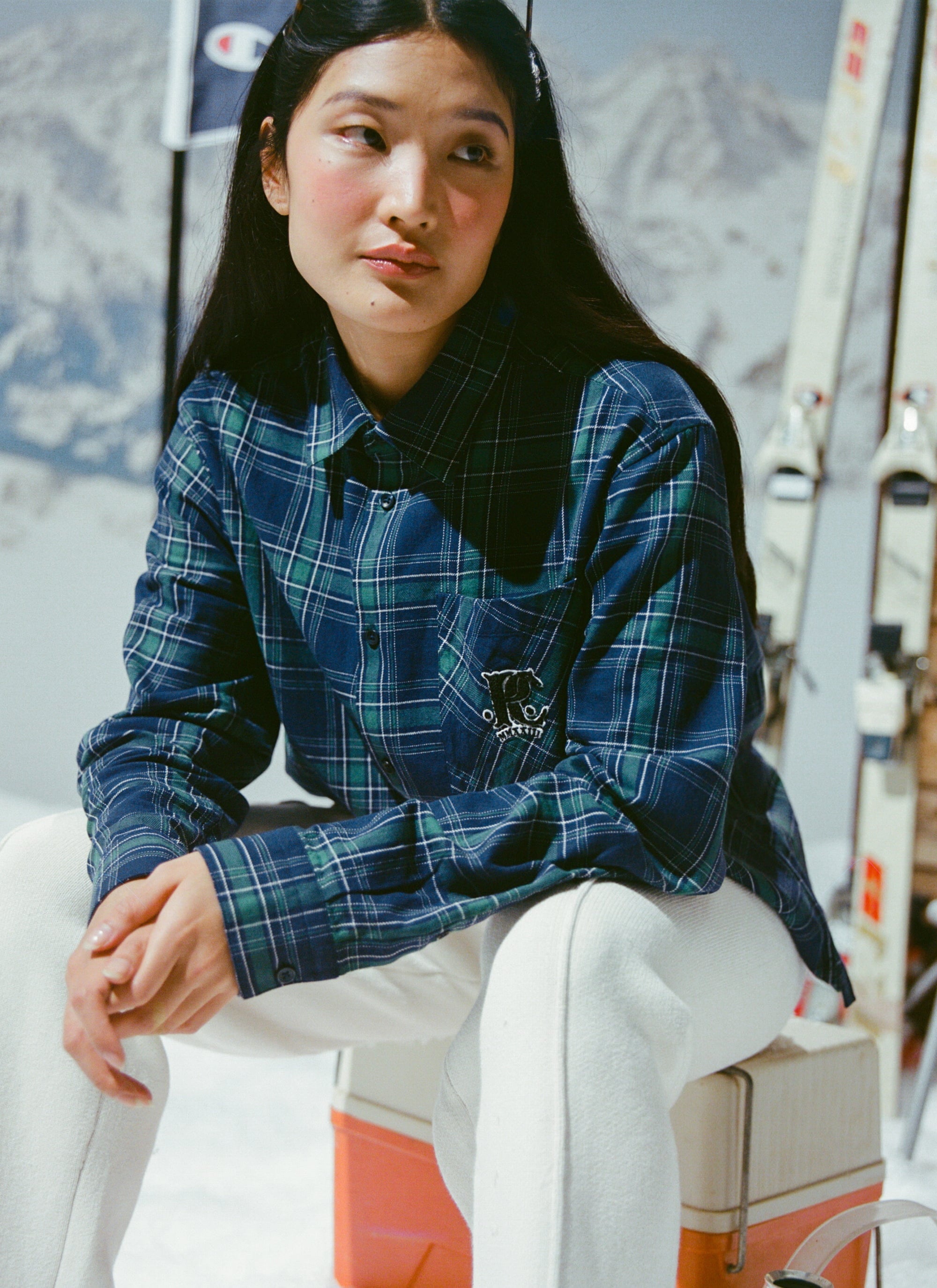 A woman models the Percival x Champion Check Boxy Long Sleeve Shirt in a blue and green tartan against a ski backdrop.