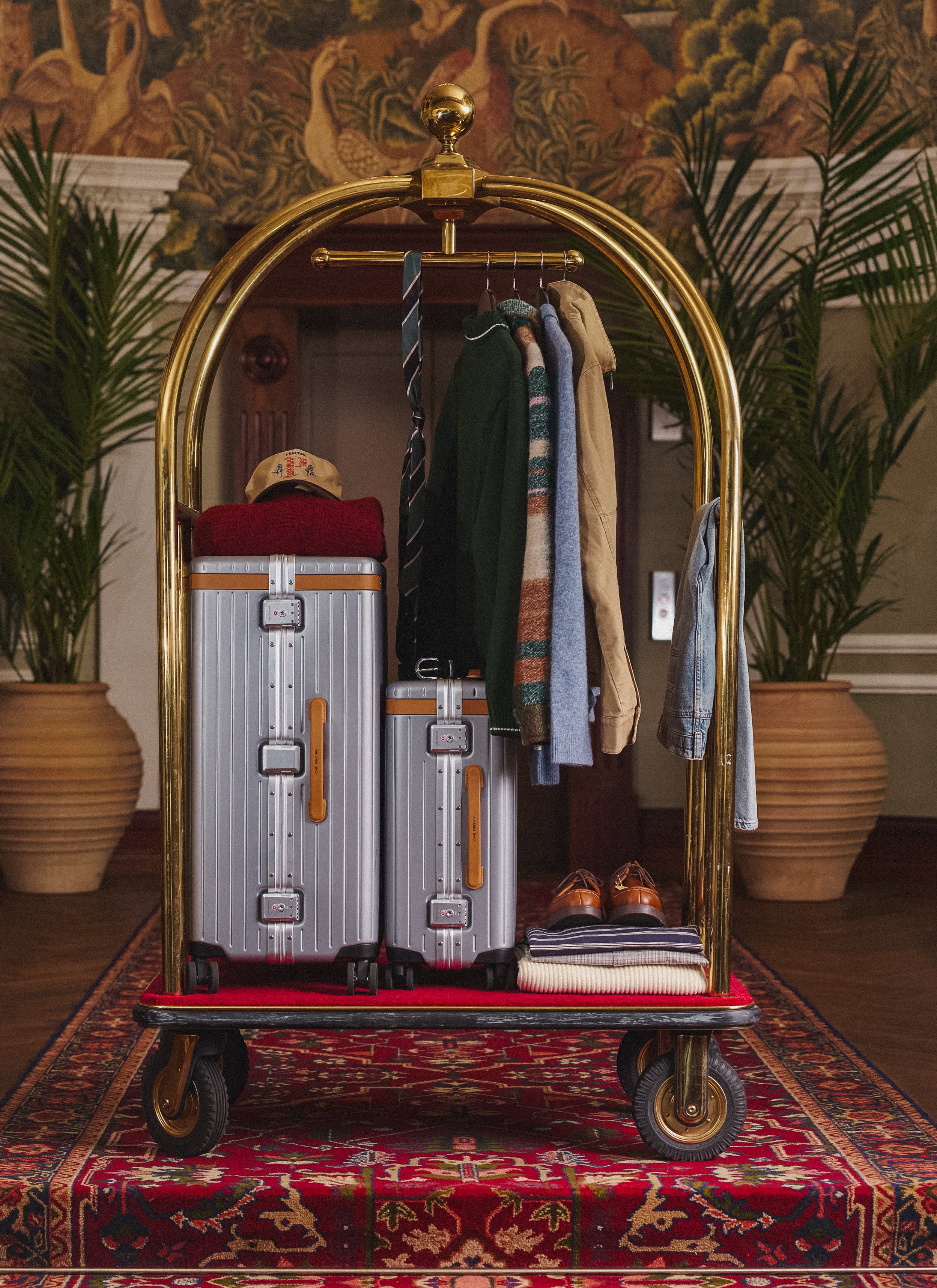 Percival AW26 collection clothing and accessories displayed on a luxury hotel luggage cart with silver suitcases.