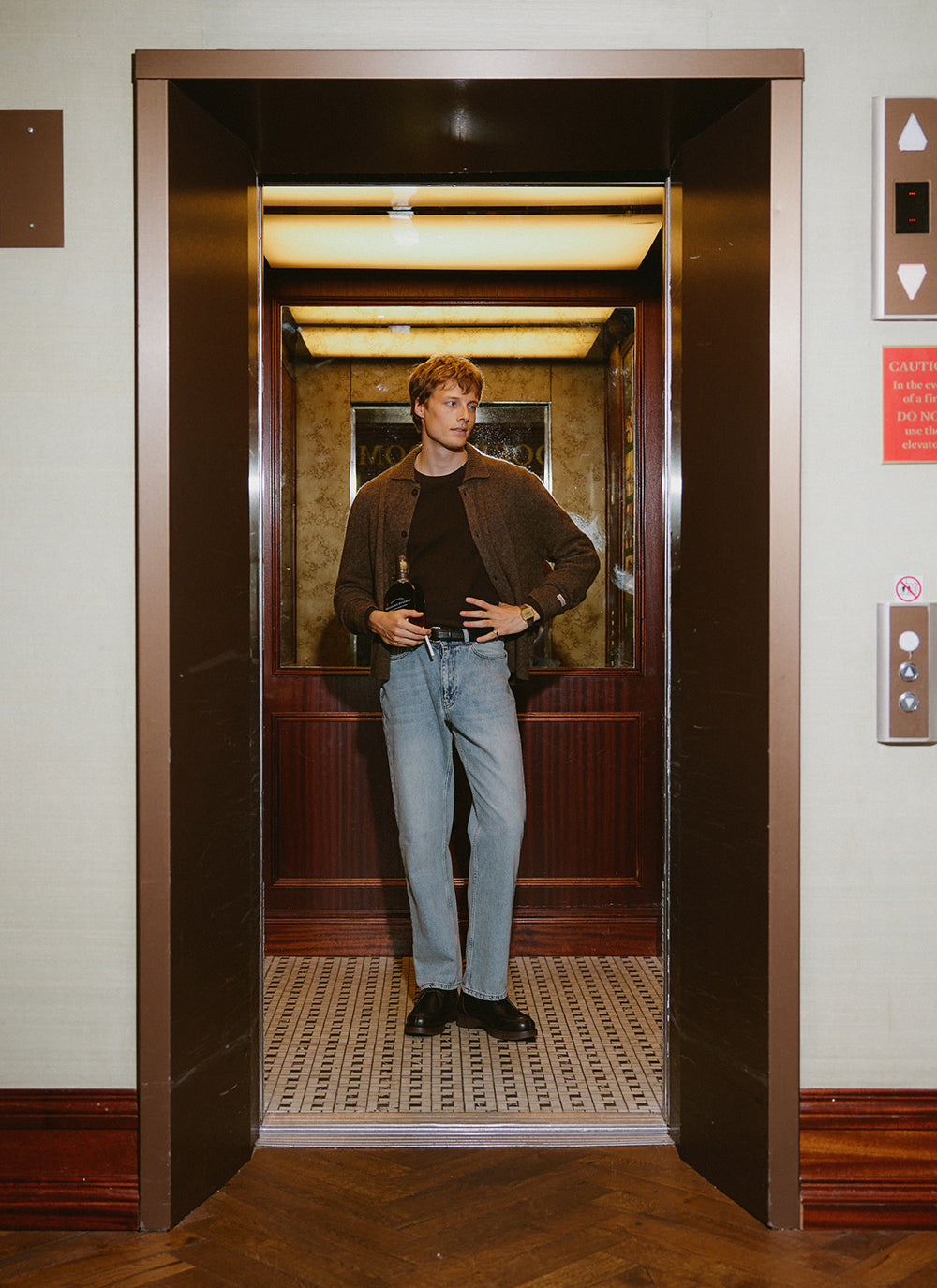 A model wears the brown Percival cardigan inside an elevator, styled with light blue jeans and dark shoes.
