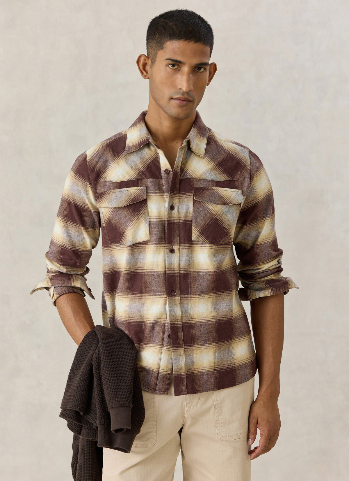 A model wearing the Percival brown checked cotton western shirt, holding a dark knit garment against a neutral backdrop.