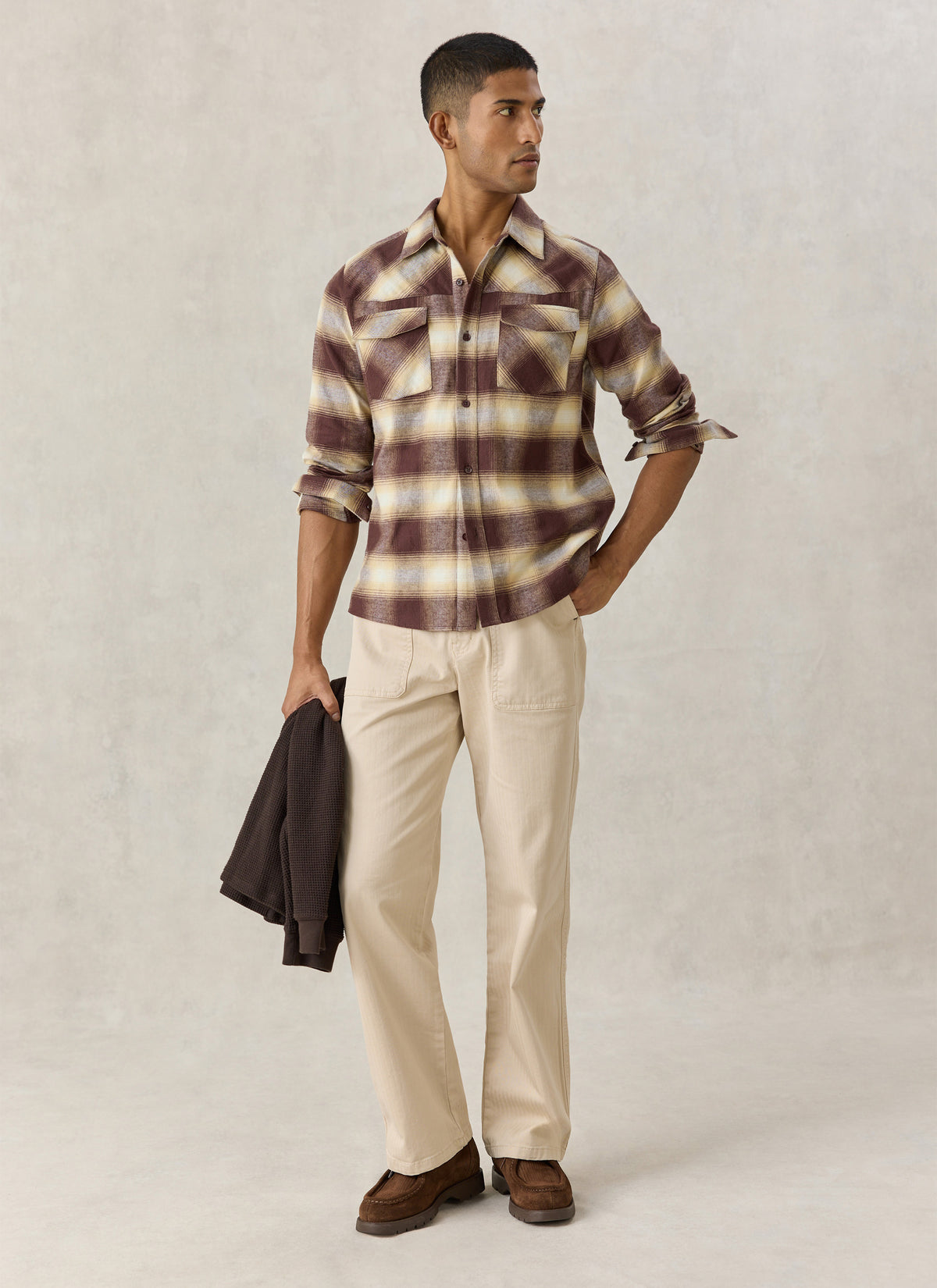 A model wearing the Percival brown checked Ashe cotton western shirt paired with light trousers and holding a garment.