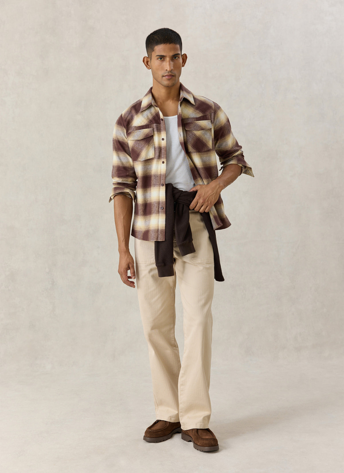 A model wearing the Percival brown checked Ashe western shirt open over a white tee, light trousers, and brown shoes.