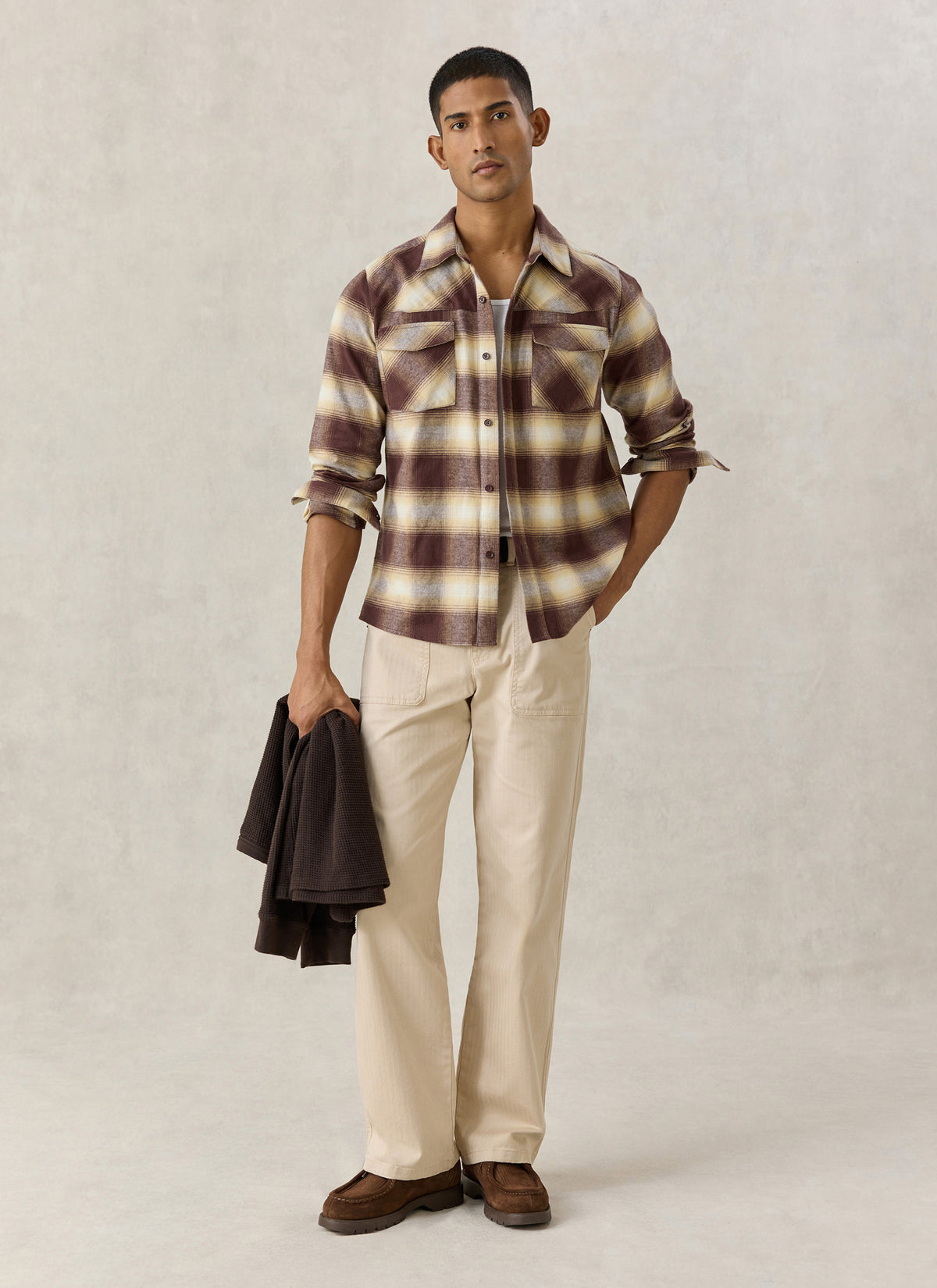 A model wearing the Percival brown checked western shirt with light trousers and loafers, holding a garment.
