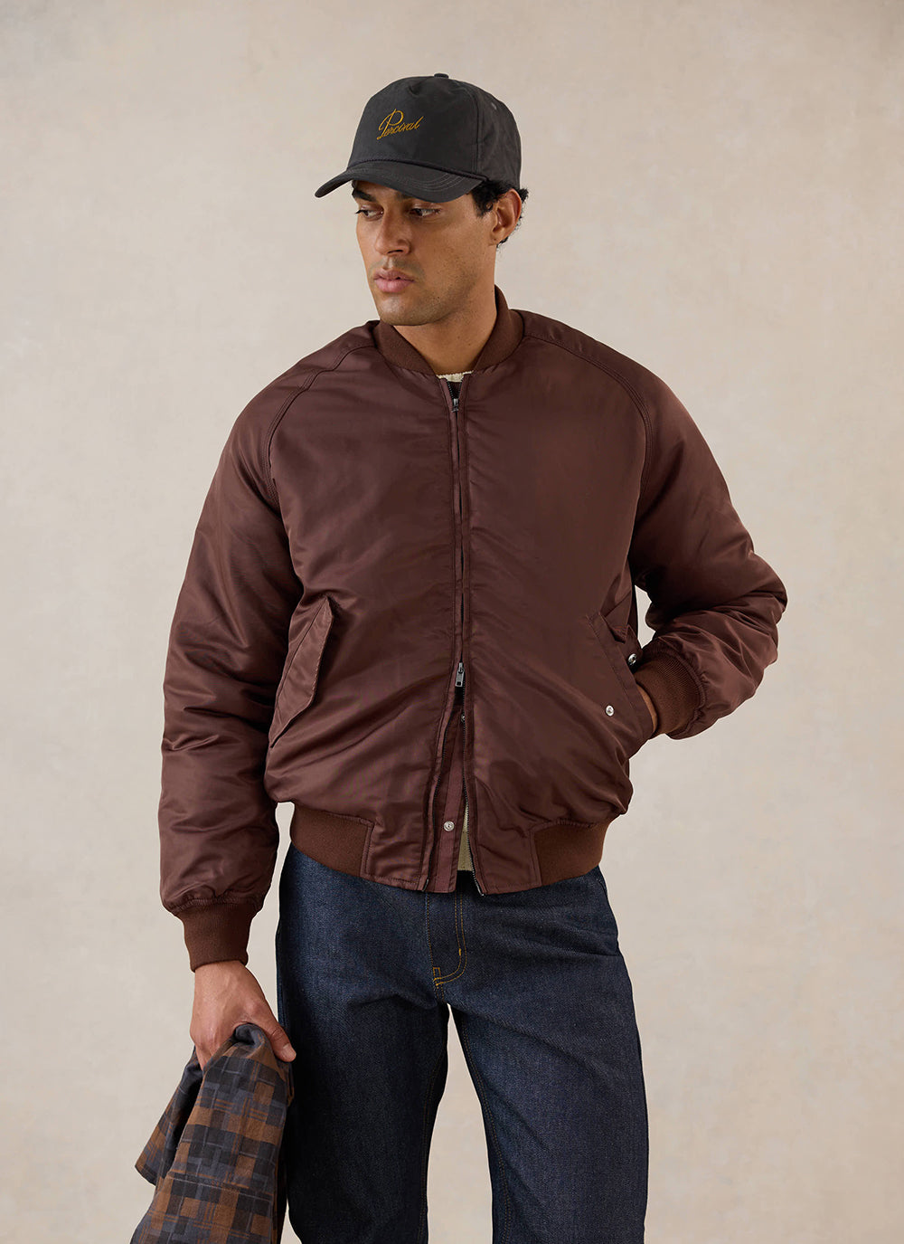 A model wears the espresso nylon bomber jacket from Percival with dark jeans and a cap in a studio.