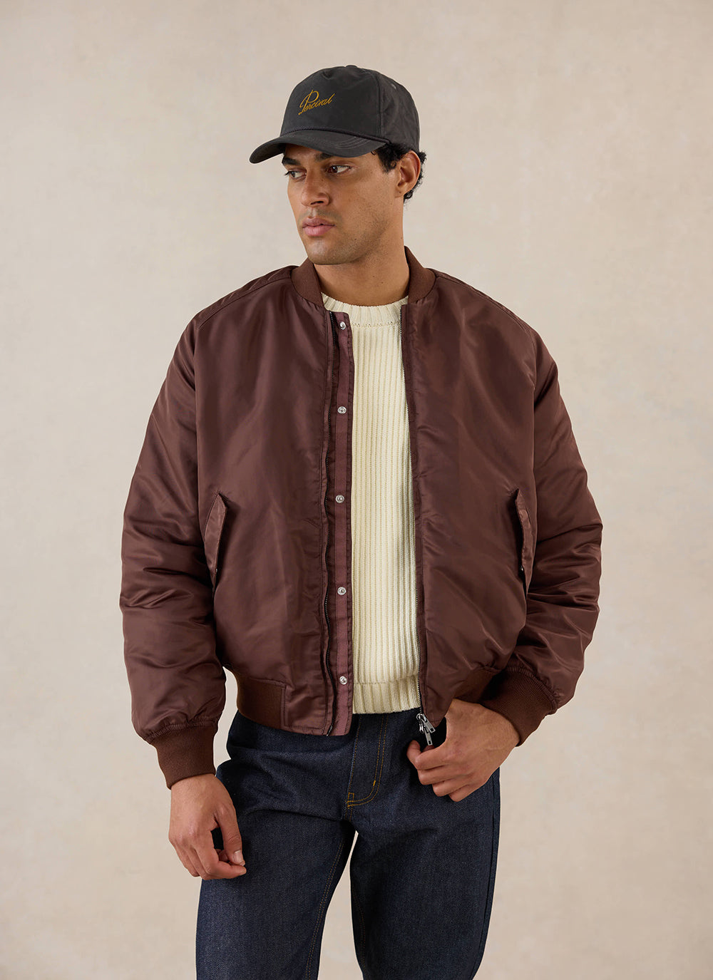 A model wears the Percival espresso nylon bomber jacket over a cream knit jumper and dark trousers in a studio shot.