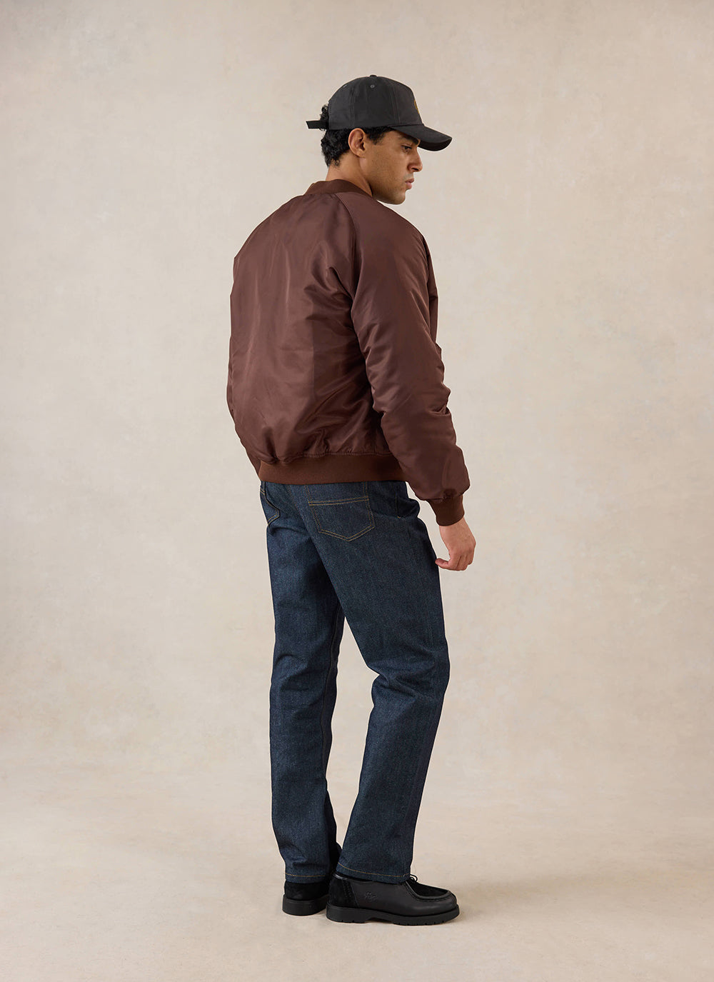 A model seen from behind wearing the espresso nylon bomber jacket from Percival with dark jeans and a cap in a studio.