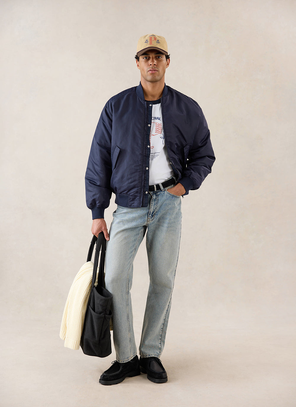 A model wears the midnight nylon bomber jacket from Percival over a white t-shirt, light jeans, and a cap in a studio.