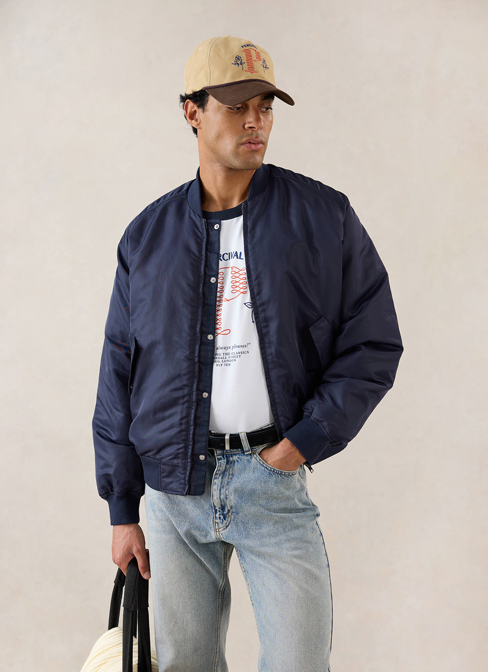 A model wears the midnight blue Percival nylon bomber jacket with a white t-shirt, jeans, and a cap in a studio.