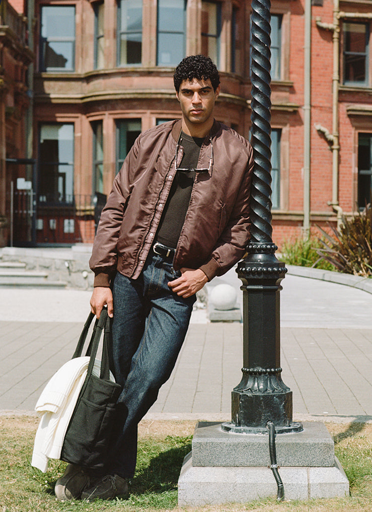 A model wears the Percival espresso brown nylon bomber jacket with a black waffle knit and raw denim jeans outdoors.