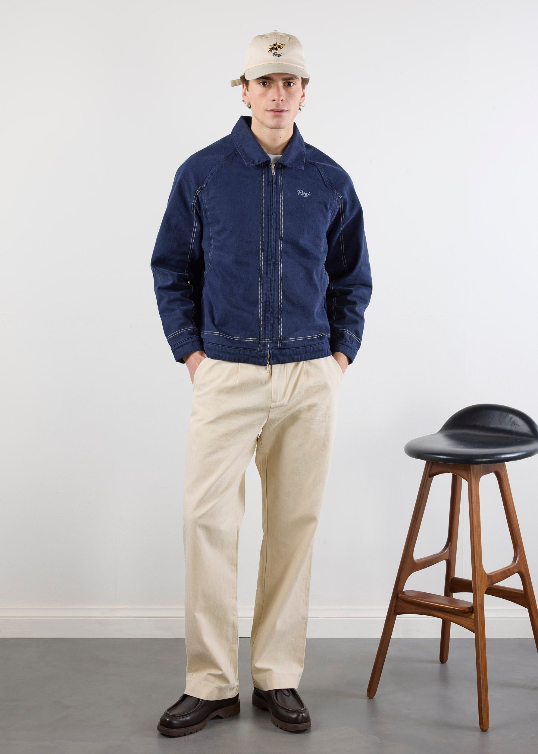 A model wearing the navy Percival Calabria Harrington Jacket with contrast stitching, styled with cream trousers and a cap.