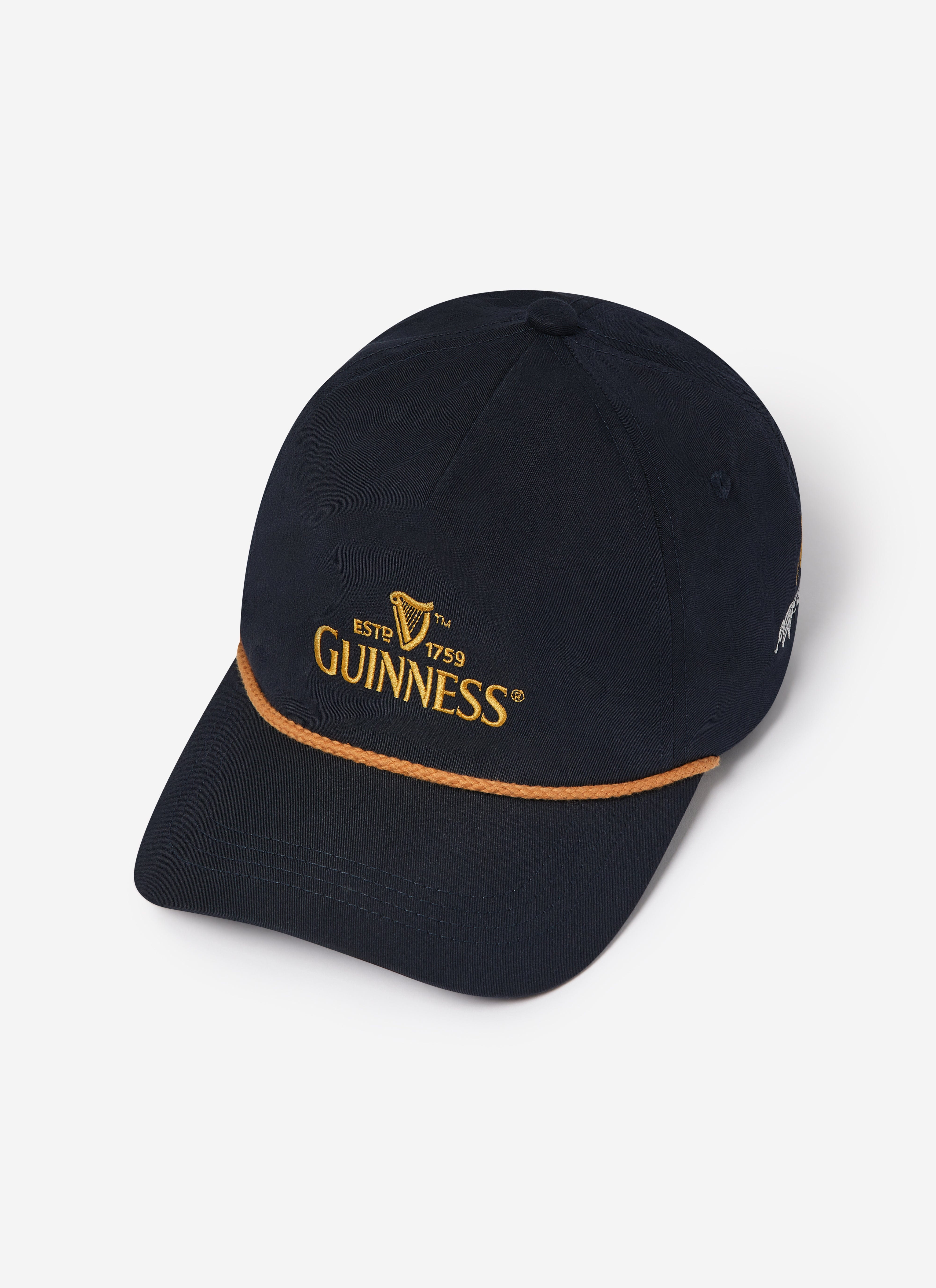 The navy Guinness x Percival cap, showing the gold embroidered Guinness logo and tan braided cord detail across the peak.