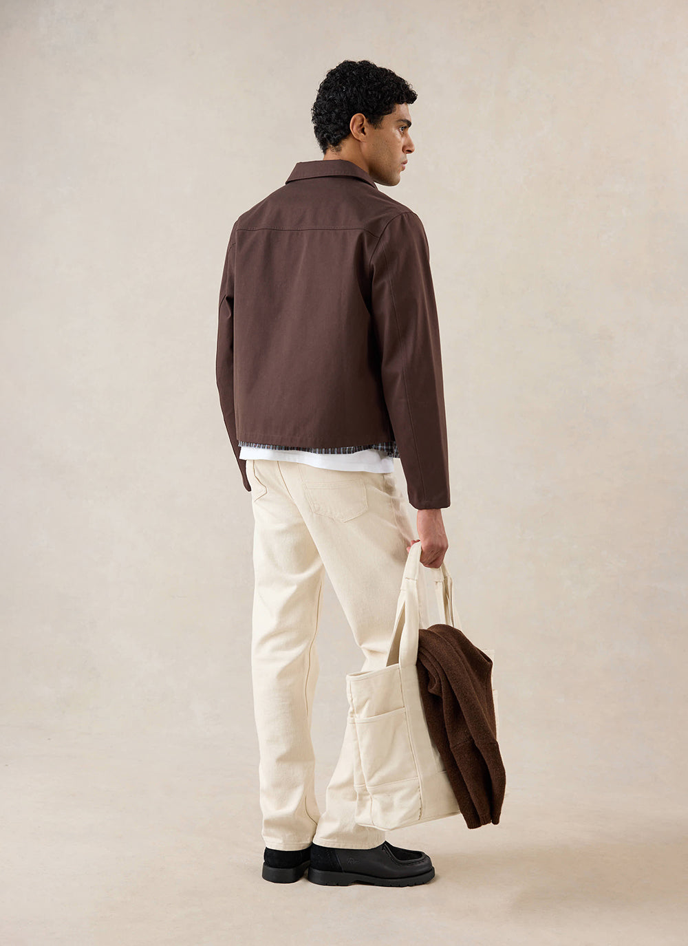A model from behind wearing the espresso Percival Carter Cropped Jacket with cream trousers and holding a tote bag.