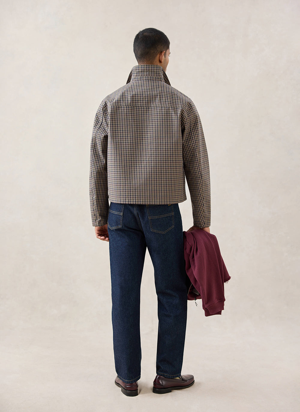 A rear-view shot of a model wearing the Watcher Check Percival Carter Cropped Trench Coat, styled with dark denim jeans.