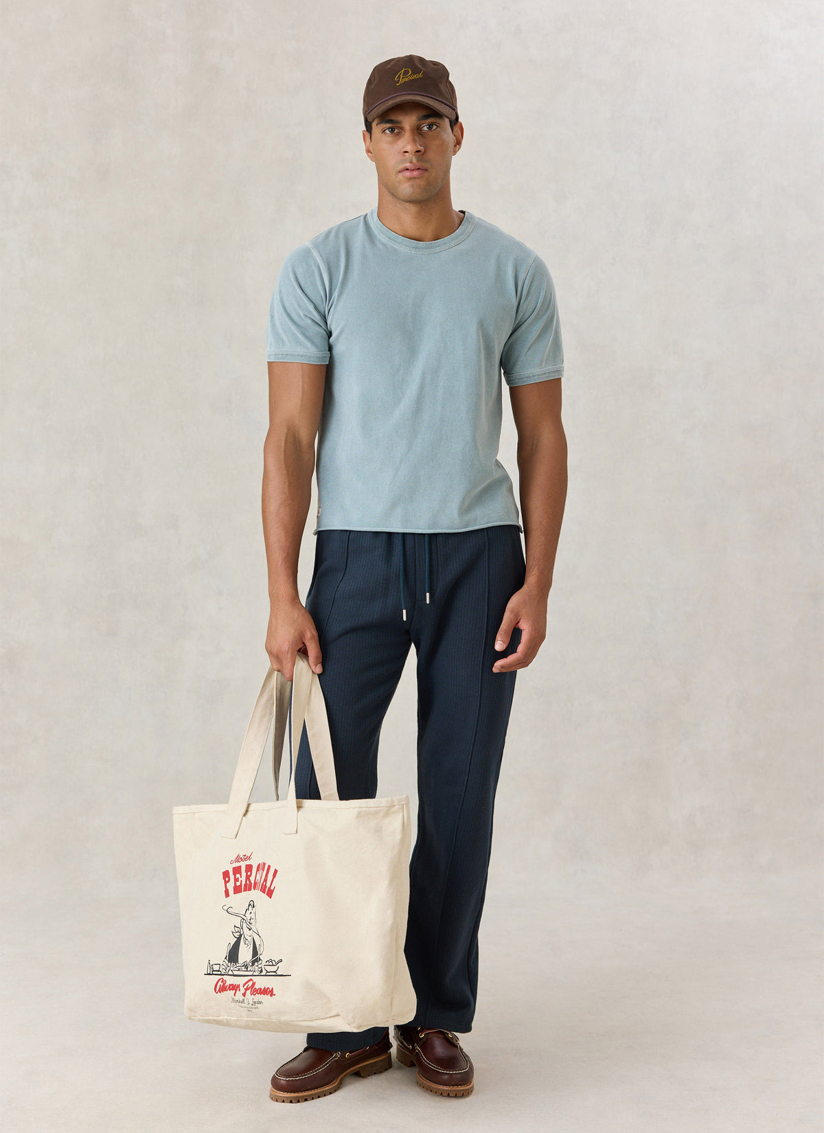 A model wearing a light blue Percival vintage-wash cropped T-shirt, dark trousers, a brown cap, and holding a tote bag.