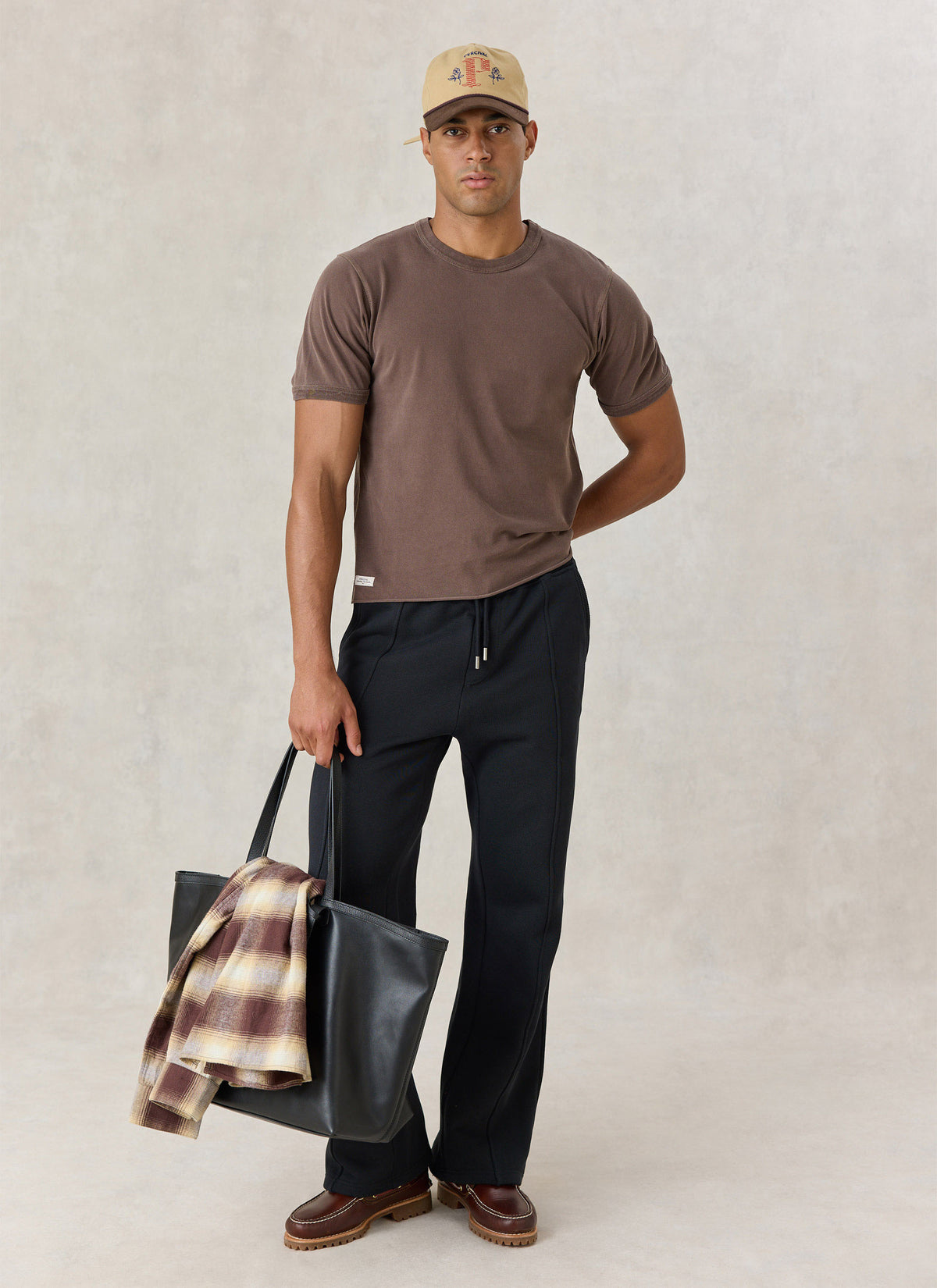 A model wearing the Percival brown vintage wash cropped T-shirt with black trousers, a cap, and holding a tote bag.