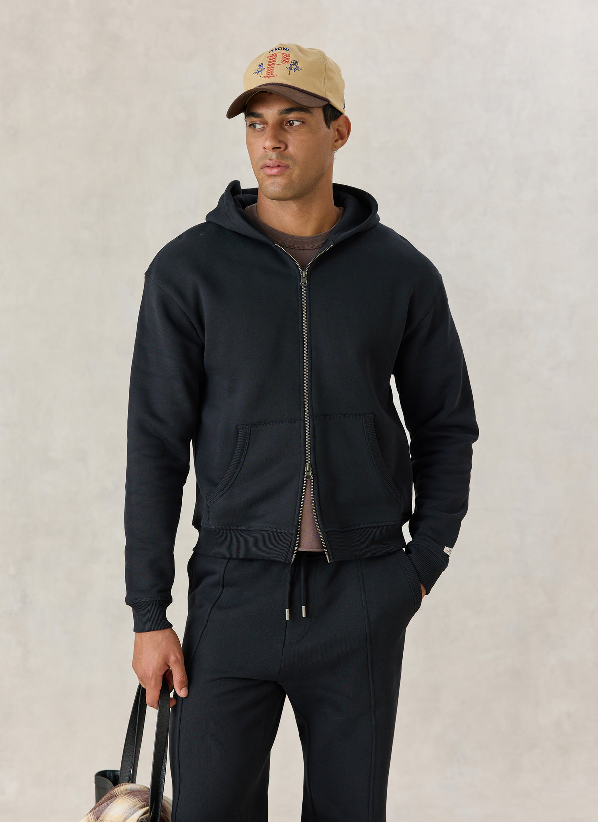 A model wearing the black Percival Chronicle zip hoodie in cotton jersey with a cap and matching trackpants.