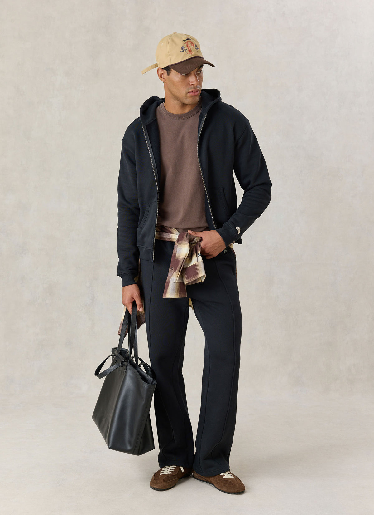 Model wearing the Percival black cotton Chronicle zip hoodie with a brown tee, black trackpants, cap, and a leather tote bag.