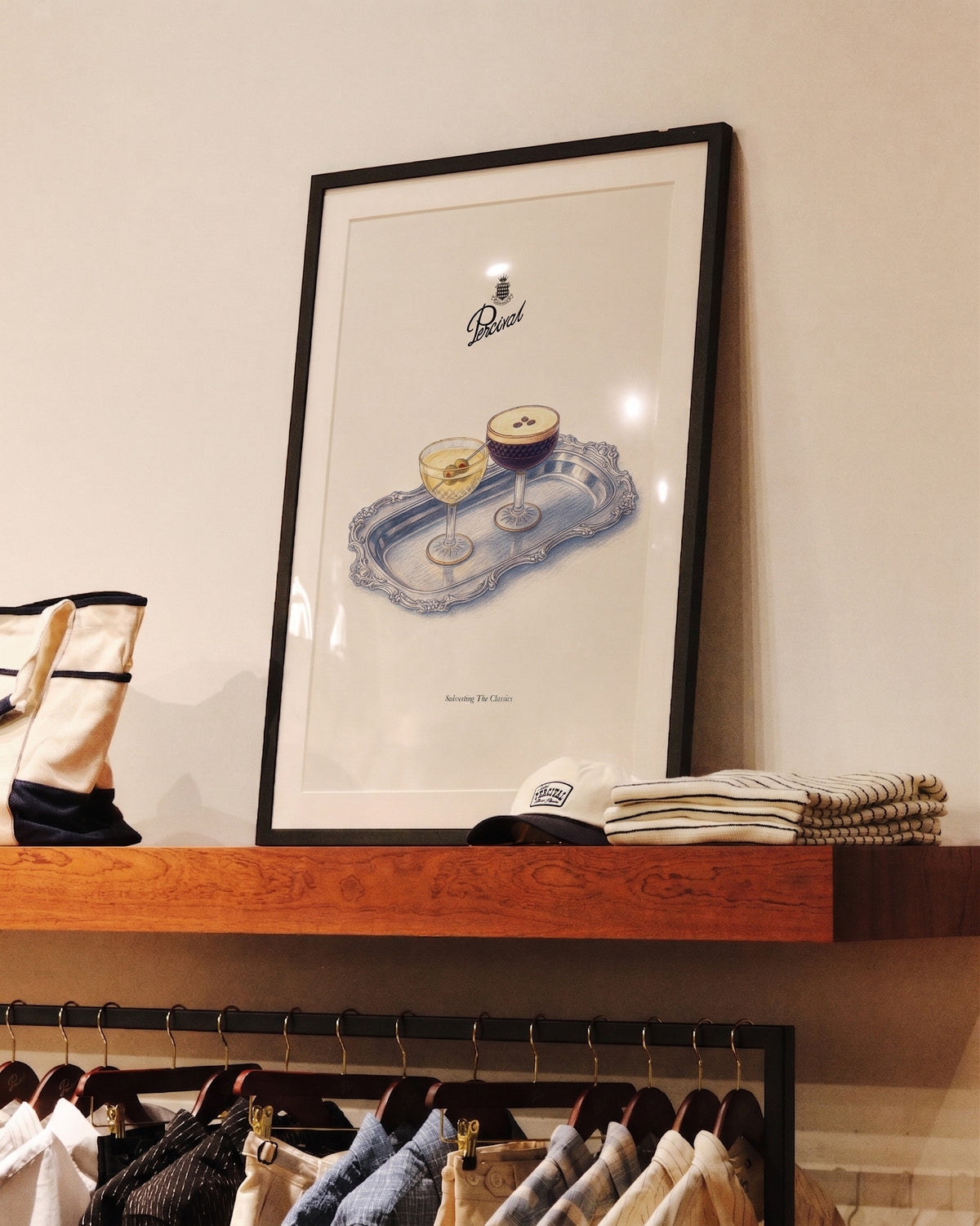 The framed Percival Cocktails Print displayed on a wooden shelf above a clothing rack in a store.