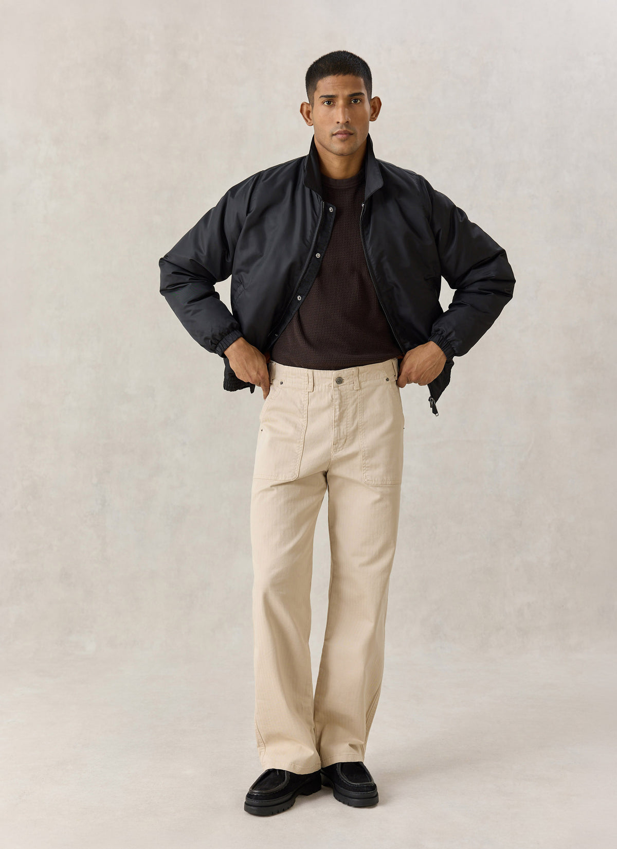 A model wearing the Percival black nylon collared bomber jacket with light trousers and a dark knit.
