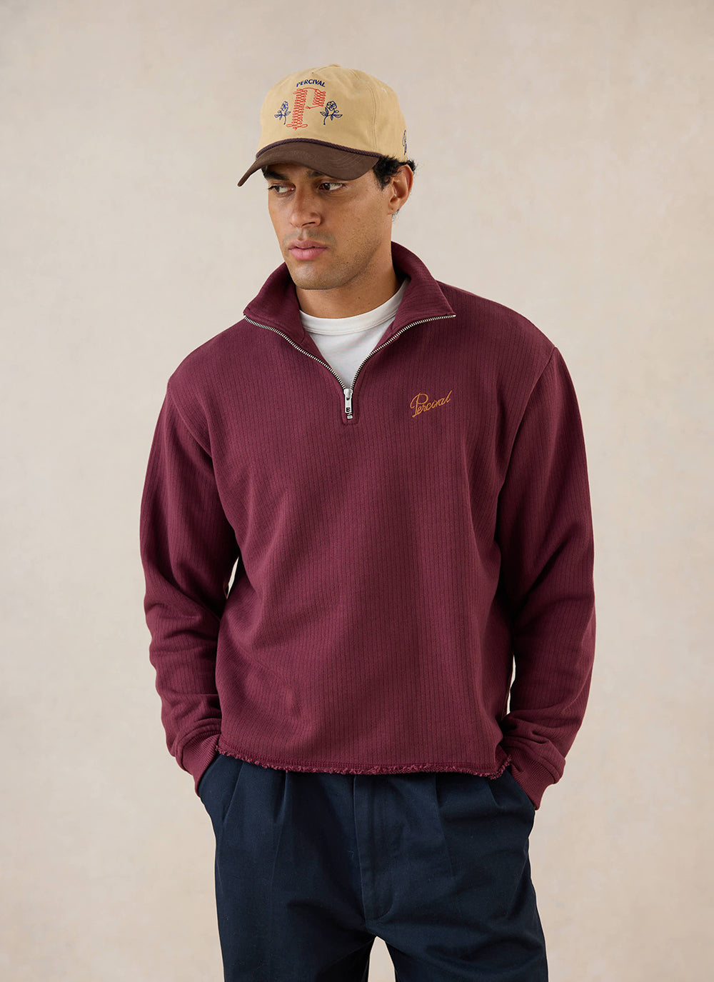 A model wears the burgundy Percival Conrad 1/4 zip funnel neck jumper, styled with a cap and dark trousers.