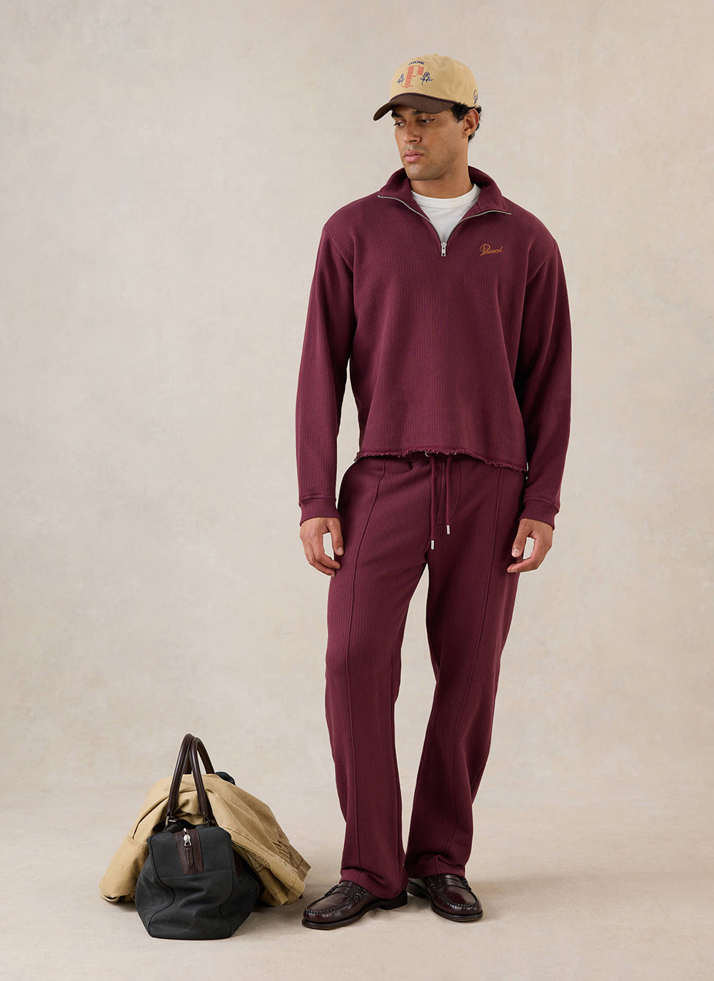 A model wears the burgundy Percival Conrad 1/4 zip jumper, styled with matching trousers, a cap, and a holdall.