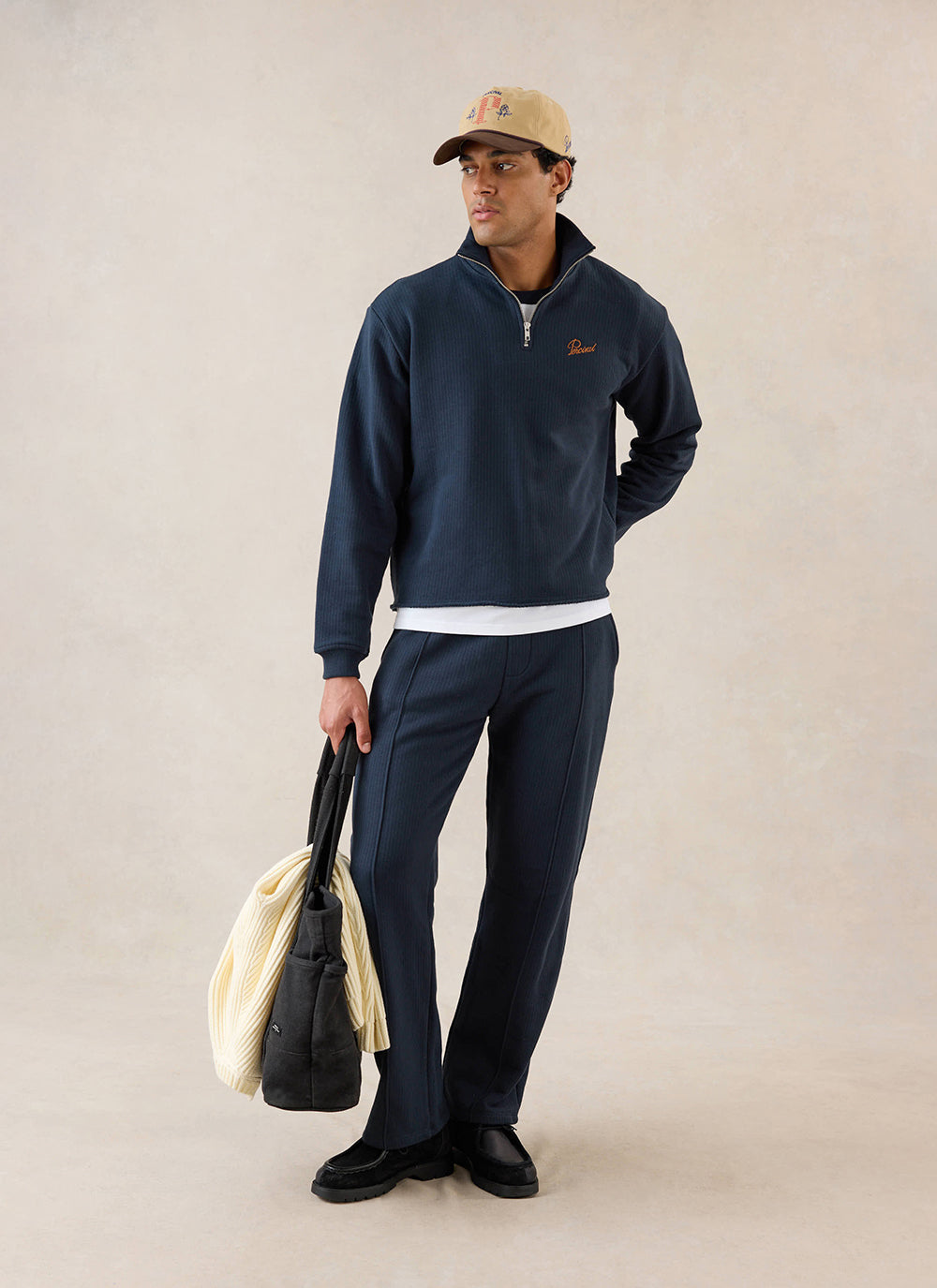 A model wears the navy Percival Conrad 1/4 zip jumper with matching trousers, a cap and a bag in a studio setting.