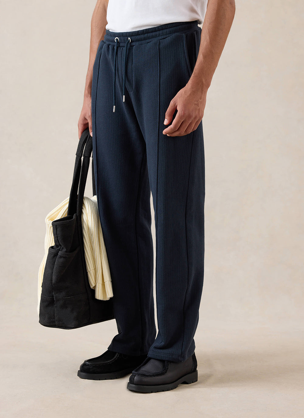 A model wears navy Percival Conrad trackpants, styled with a tote bag and black shoes in a studio shot.