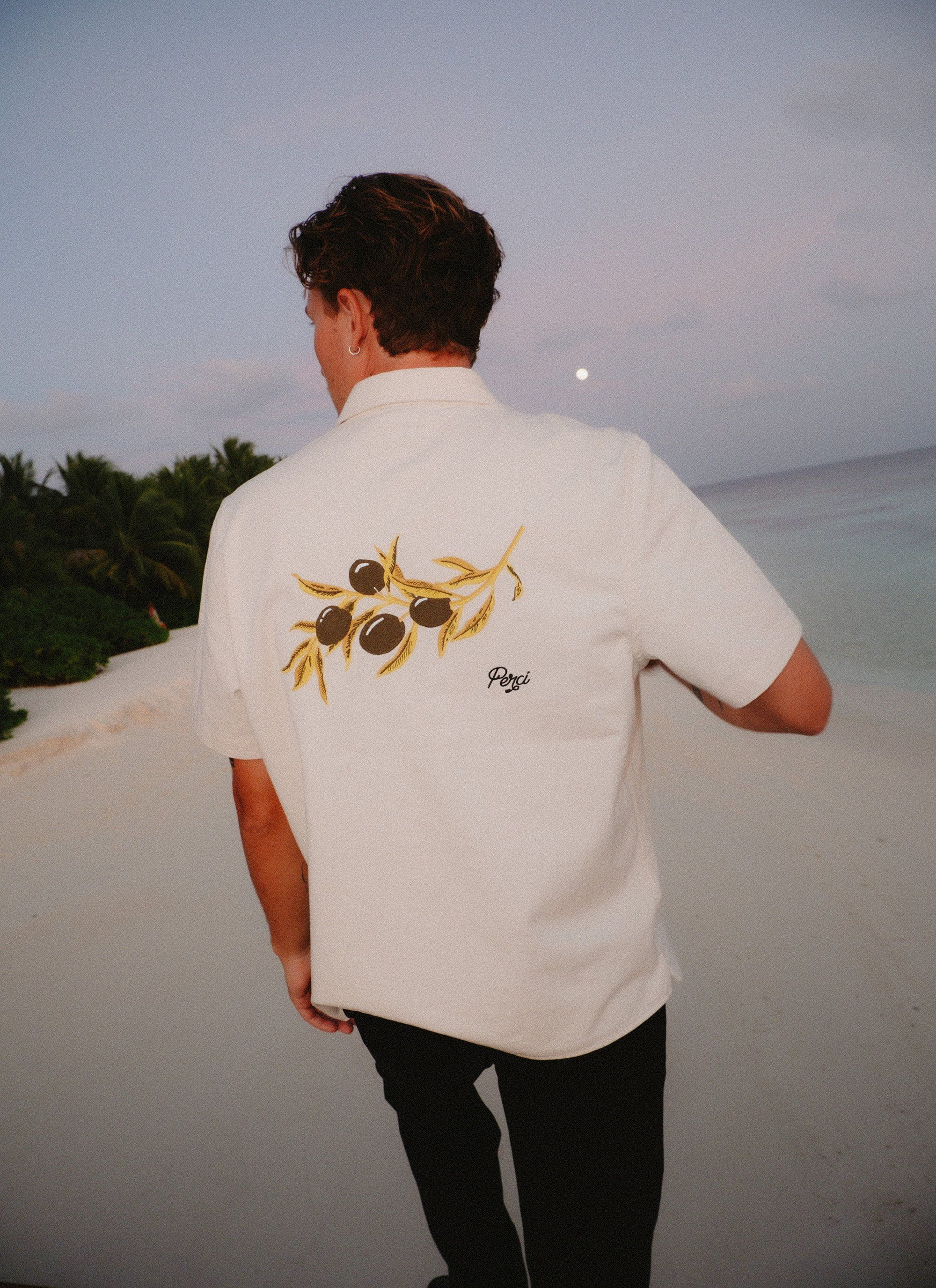 A man on a beach at dusk wearing the white Percival Olive Embroidered Cropped Shirt, showing the olive branch embroidery.