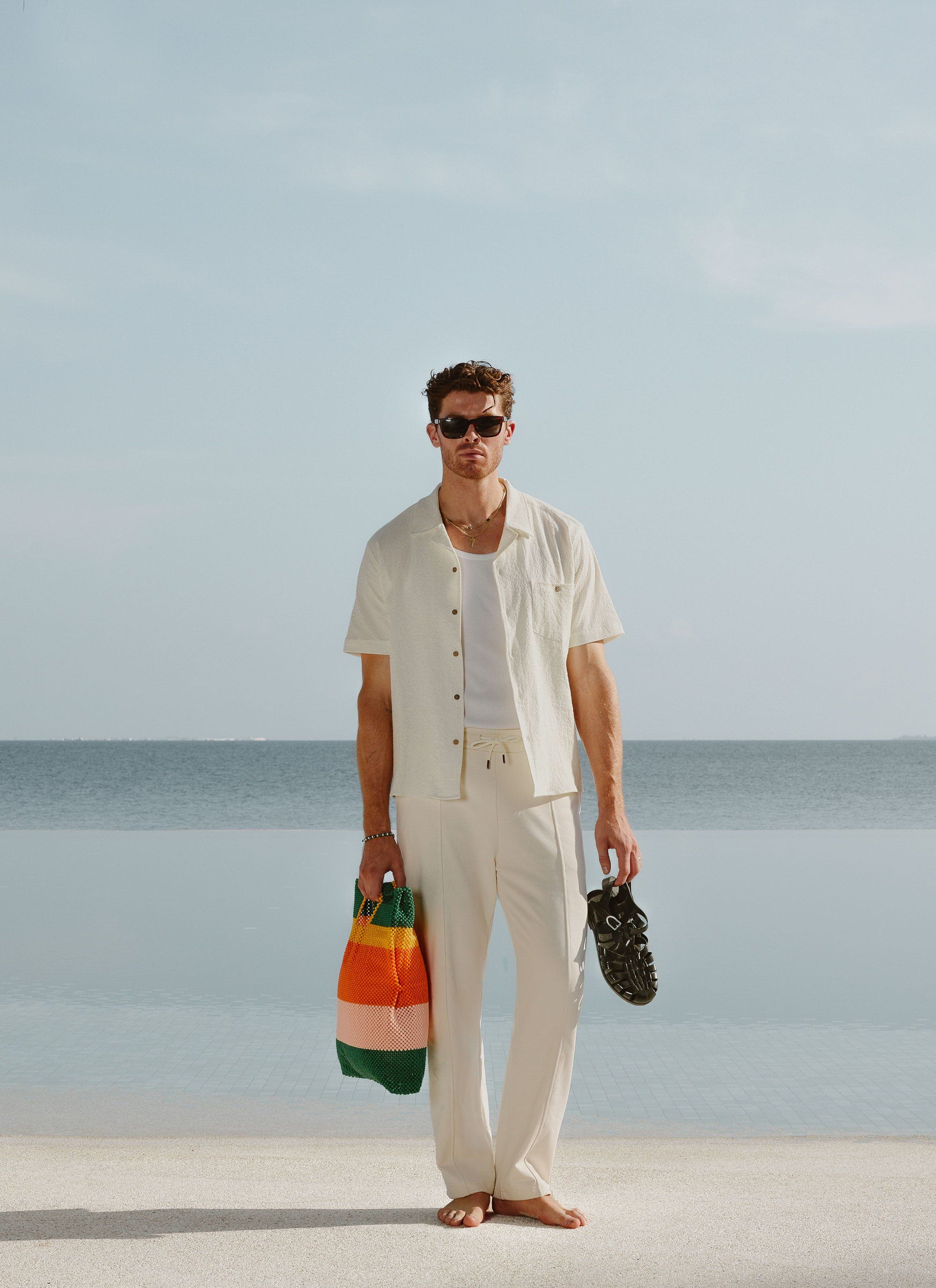 A model wears the Percival ecru seersucker Cuban shirt with matching trousers, standing by the sea.
