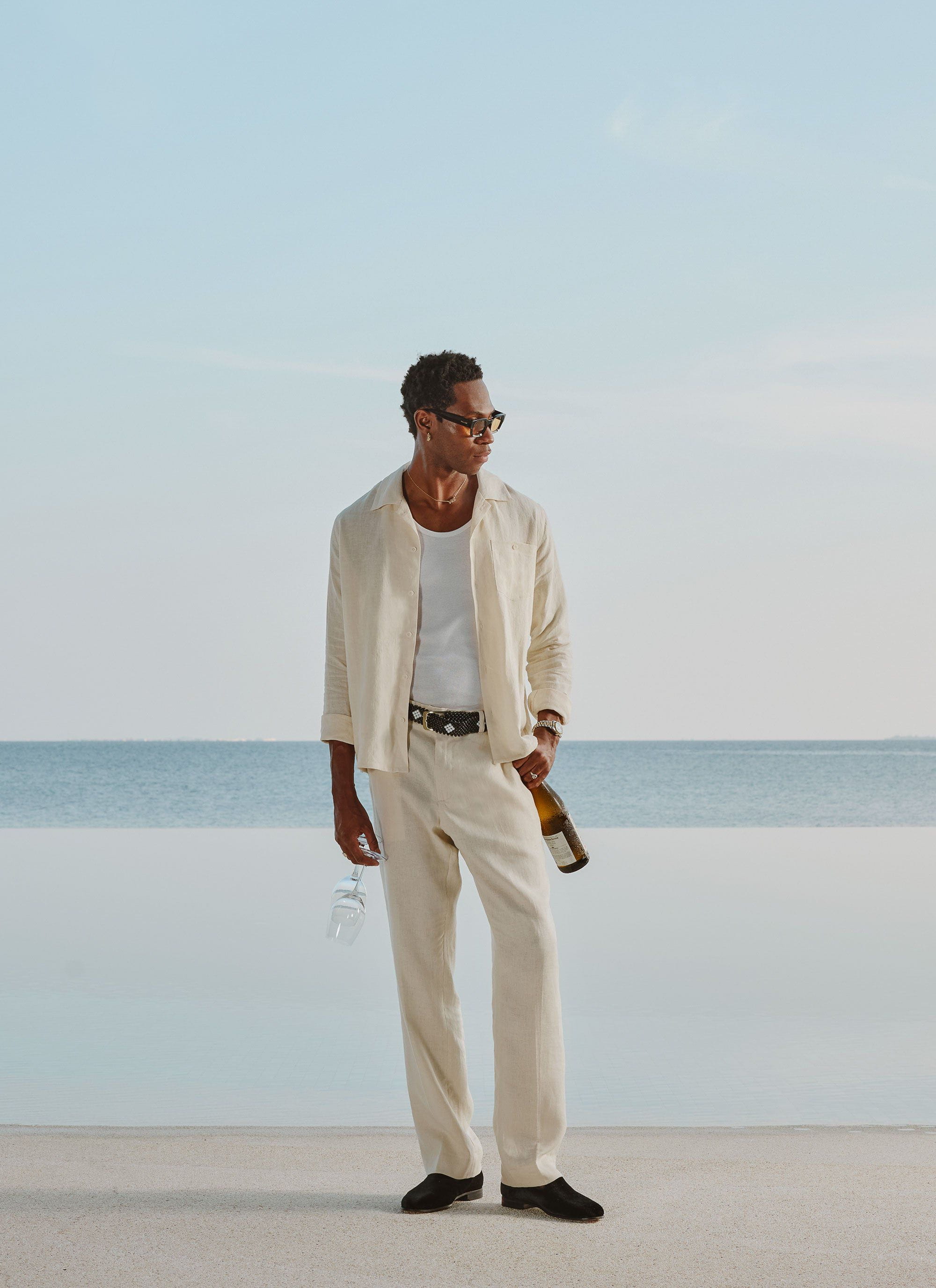 A model wears the Percival Long Sleeve Cuban Linen Shirt in cream with matching trousers, standing by the sea with wine.