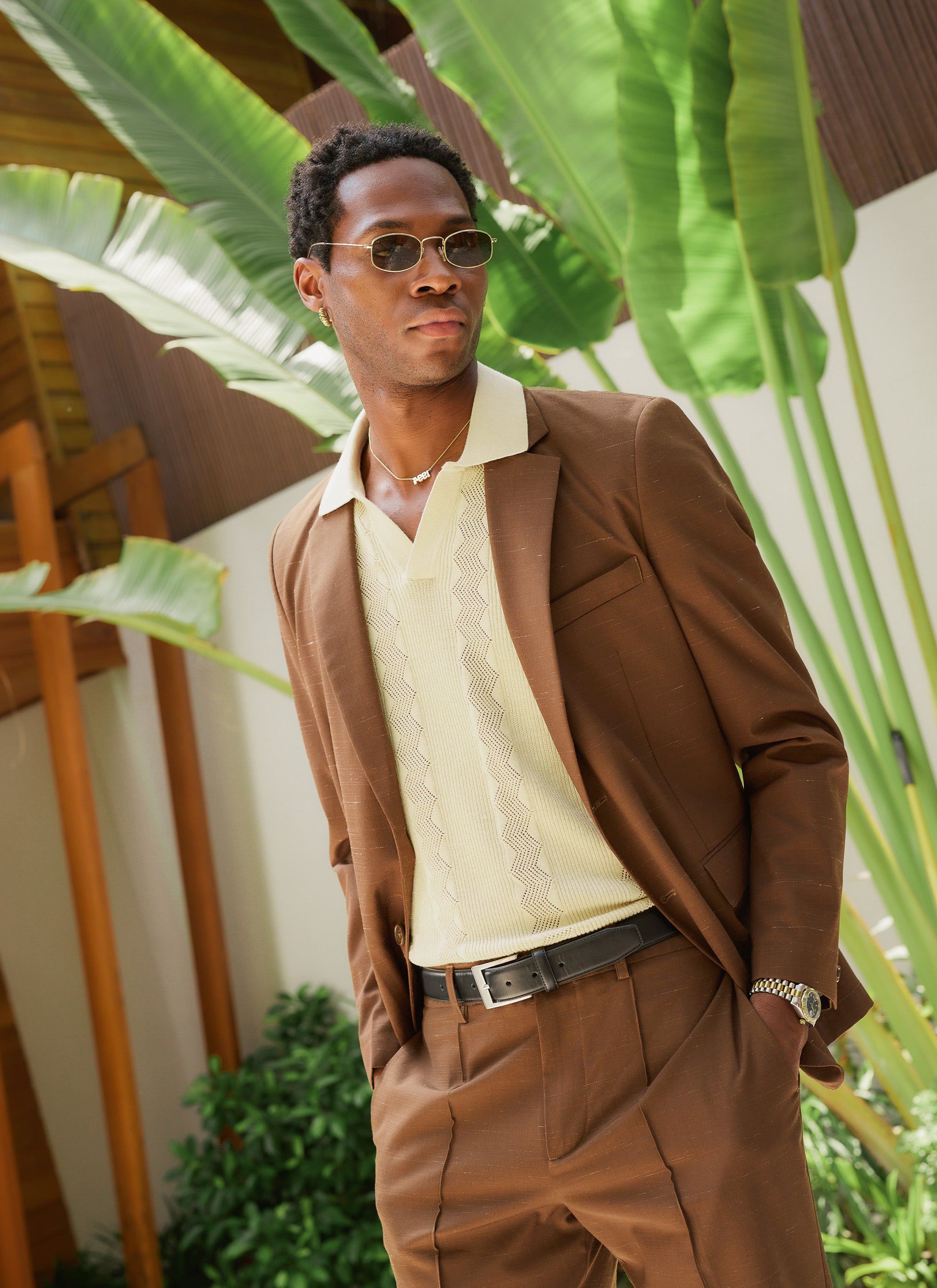 A model wears the ecru Percival Casa Martini Knitted Polo, styled under a brown suit jacket against a leafy green backdrop.