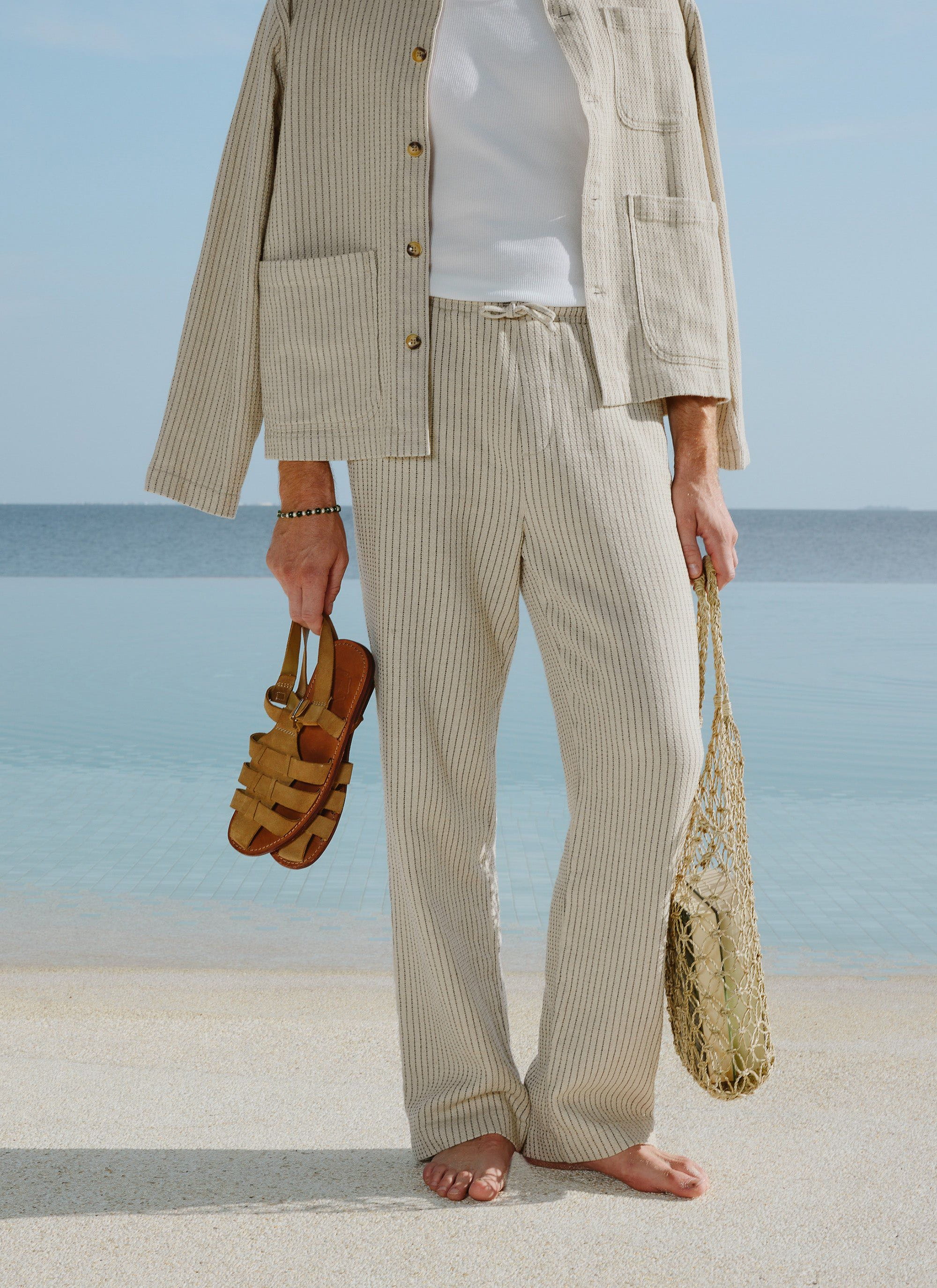 A model wears the Percival Sorrento Linen Trousers and matching jacket in ecru stripe, standing barefoot by the water.