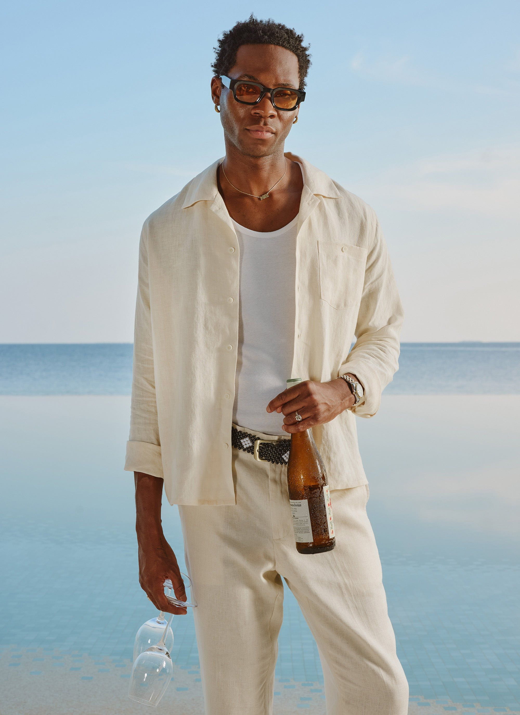 A model wears the cream Percival Long Sleeve Cuban Linen Shirt open over a vest with matching trousers by a pool.