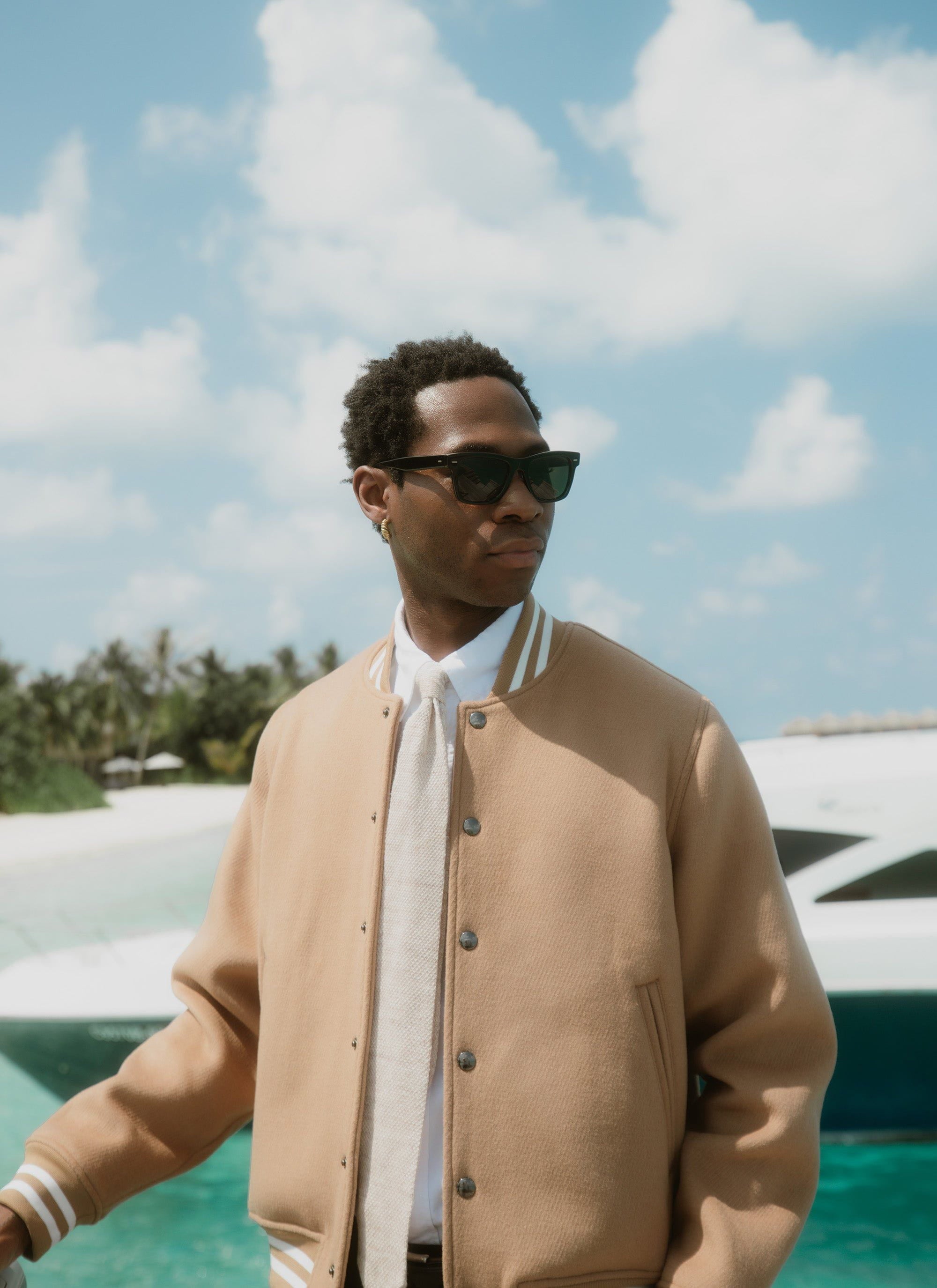 A model wears the Percival Puglia Varsity Bomber in tan wool, styled with sunglasses against a tropical sea backdrop.