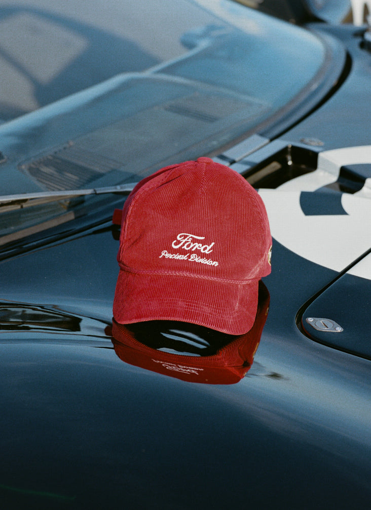 Red corduroy Ford x Percival trucker cap with white embroidery, resting on the hood of a vintage racing car.