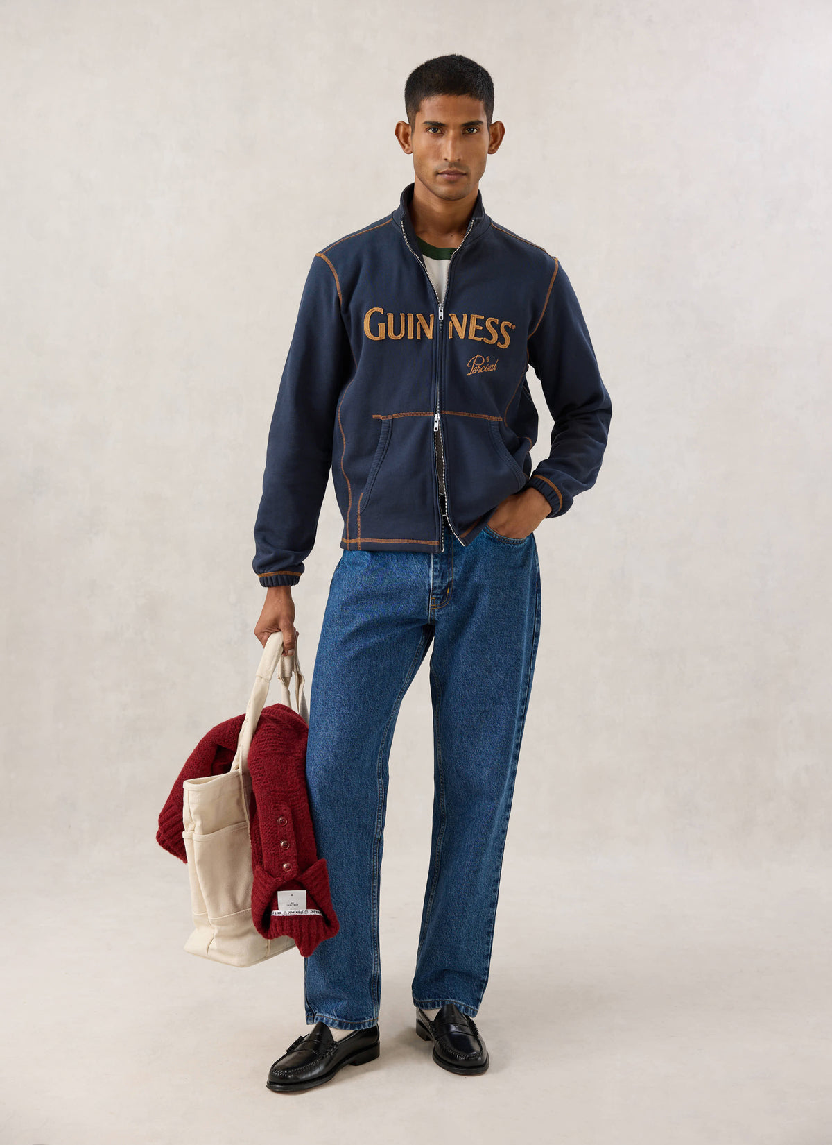 Model wearing the navy Guinness x Percival funnel neck zip up sweatshirt, blue jeans, holding a tote bag.