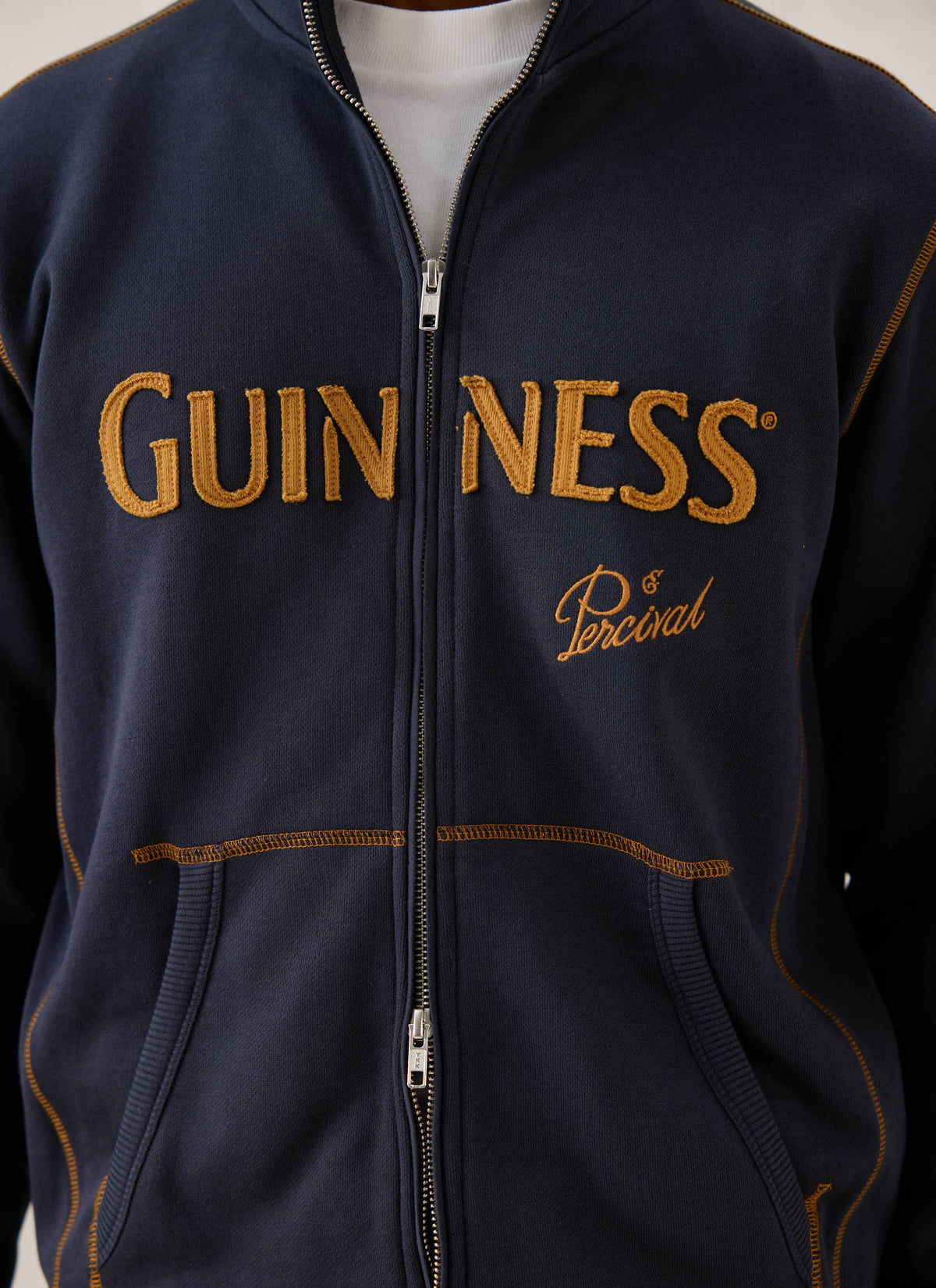 A close-up of the navy blue Guinness x Percival zip-up sweatshirt, showing the gold appliqué branding and contrast stitching.