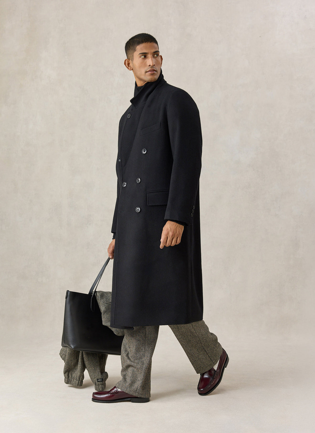A model wearing the Percival black wool blend double-breasted overcoat with patterned trousers and holding a black tote bag.