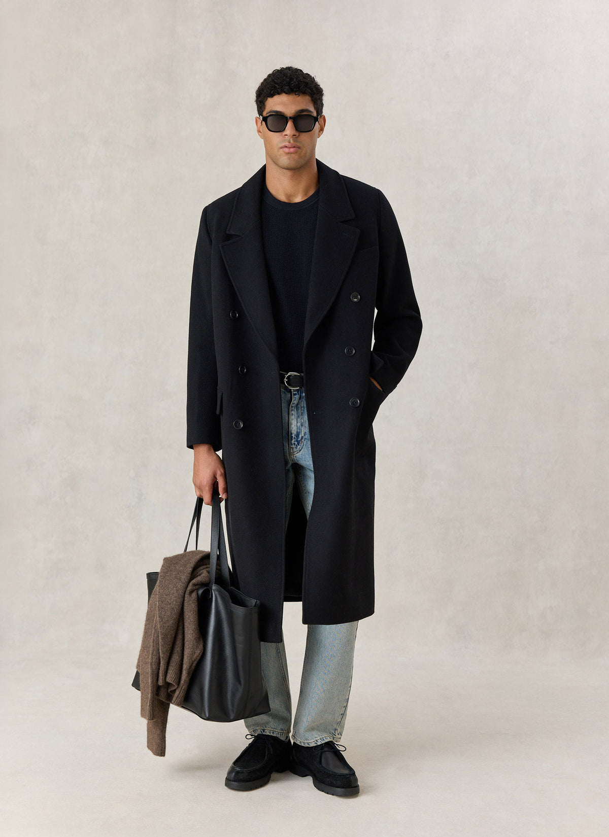 A model wearing the Percival black wool blend double-breasted overcoat with jeans, sunglasses, and a black tote bag.