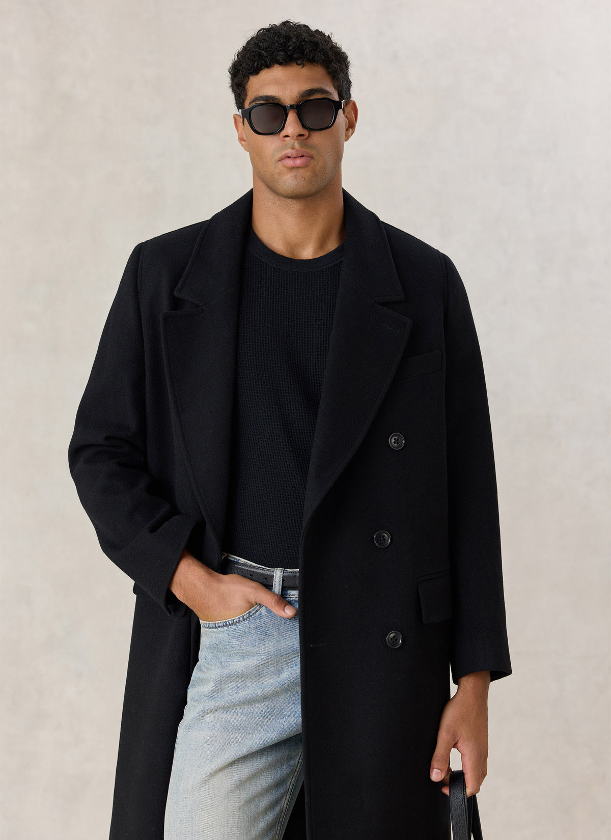 Model wearing the Percival black wool blend double-breasted overcoat over a black knit and light jeans with sunglasses.