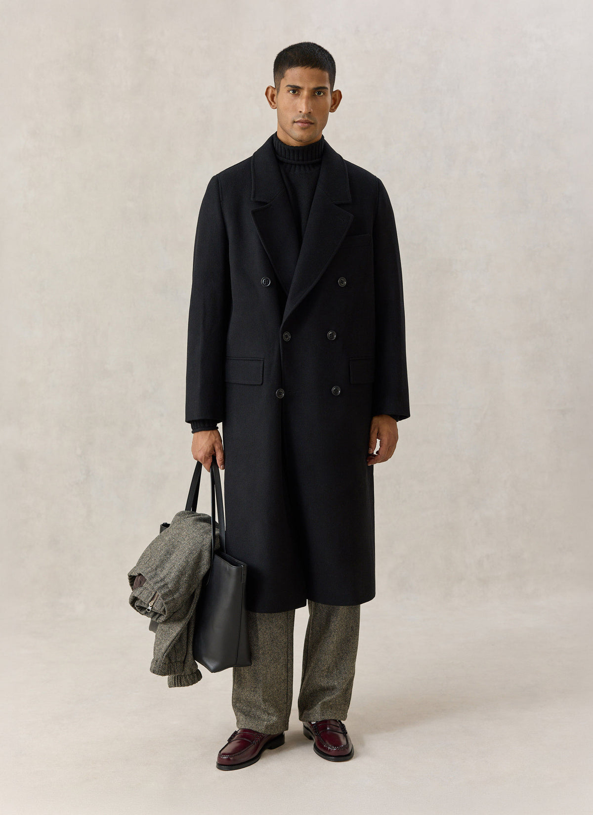 A model wearing a black wool-blend double-breasted overcoat by Percival, holding a dark leather tote and patterned trousers.