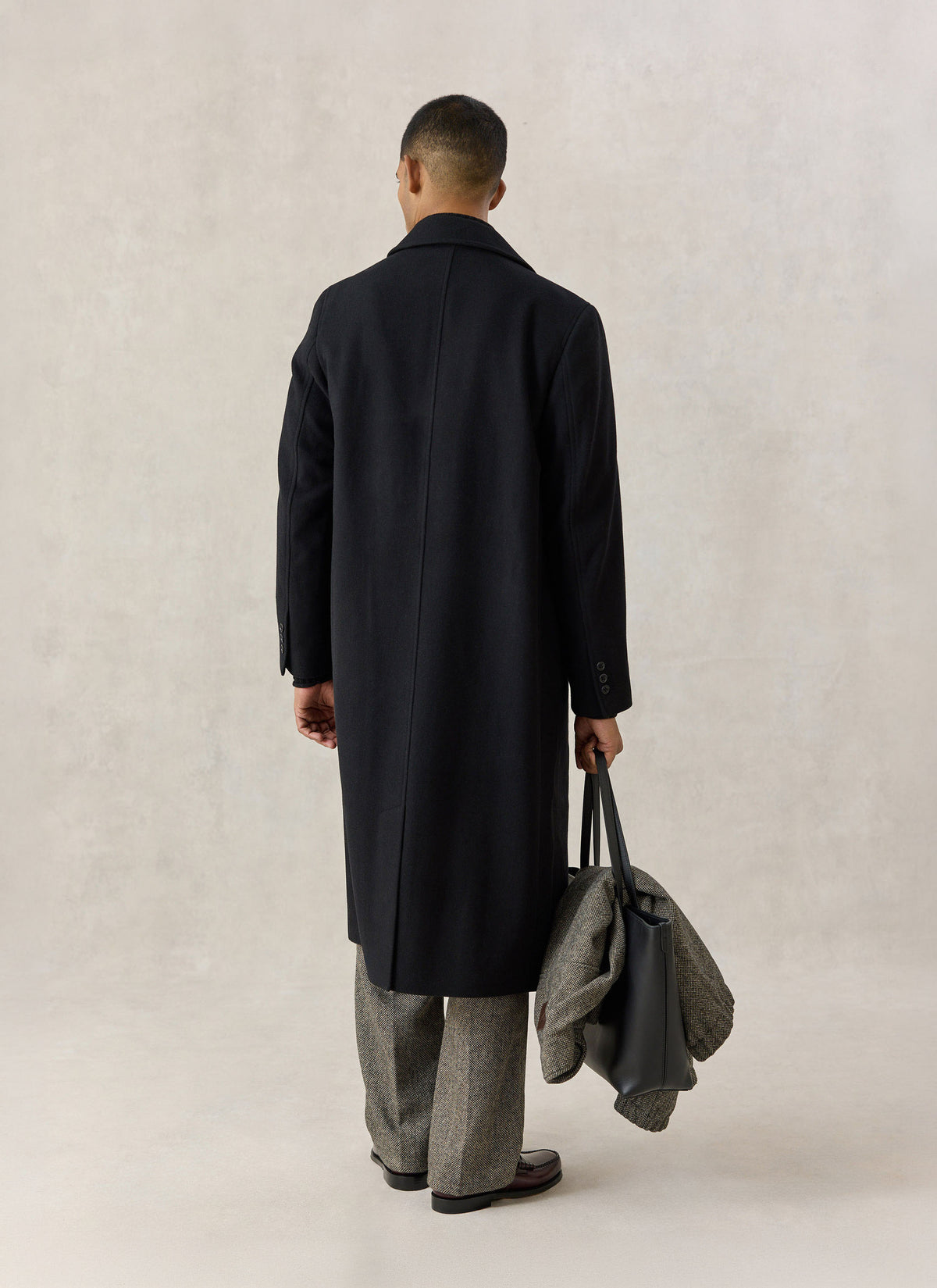 A model from the back wearing the Percival black wool overcoat and holding a black tote bag.